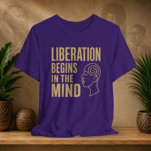 Purple t-shirt with 'Liberation Begins in the Mind' text and a face design on a wooden surface with plants and decorative items.