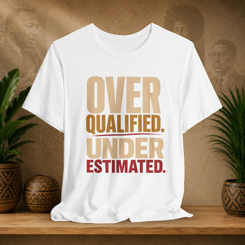 A white short-sleeve graphic T-shirt displayed on a wooden surface, with the slogan 'OVER QUALIFIED. UNDER ESTIMATED.