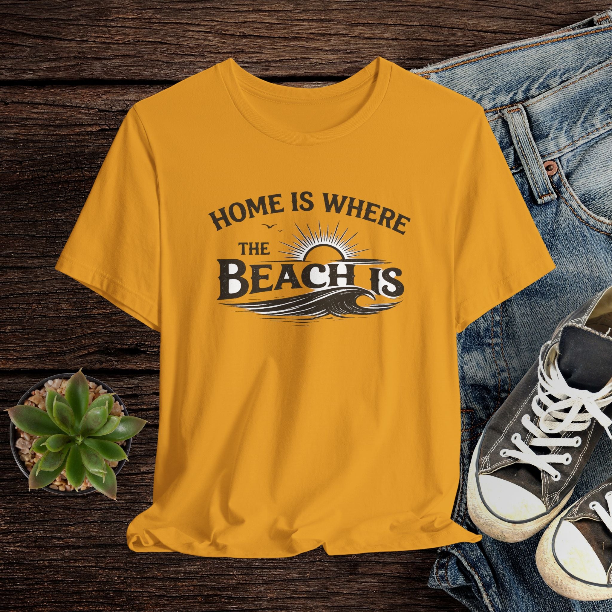 Mustard t-shirt with 'Home is where the beach is' text on a wooden surface with jeans and sneakers.