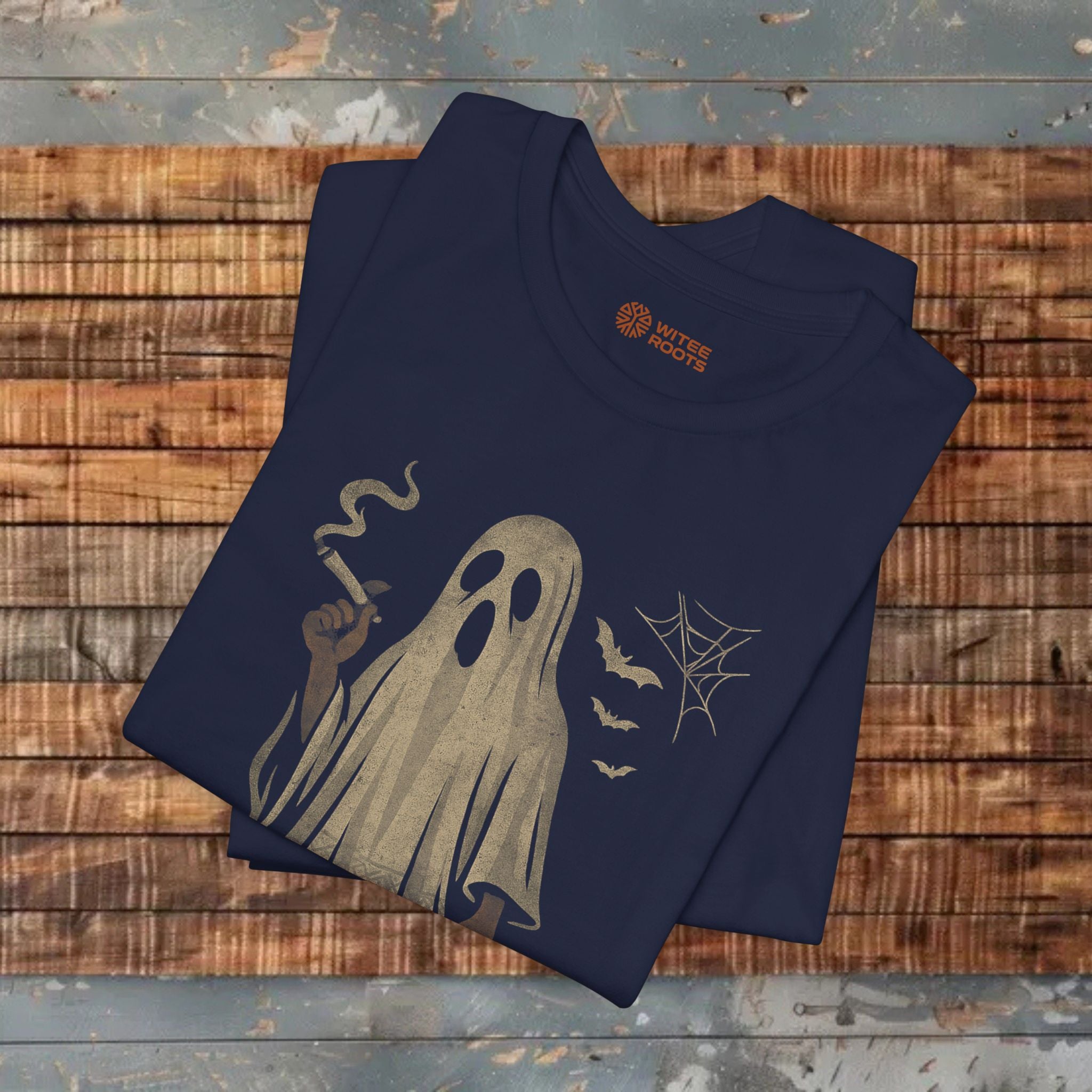 A navy blue folded t-shirt with a Halloween graphic of a smoking ghost, displayed on a rustic wooden plank surface. The shirt features a beige ghost graphic holding a cigarette, a spiderweb, three bats, and a smoke signal, with a visible 'Wild Fire Designs' logo printed inside the collar.