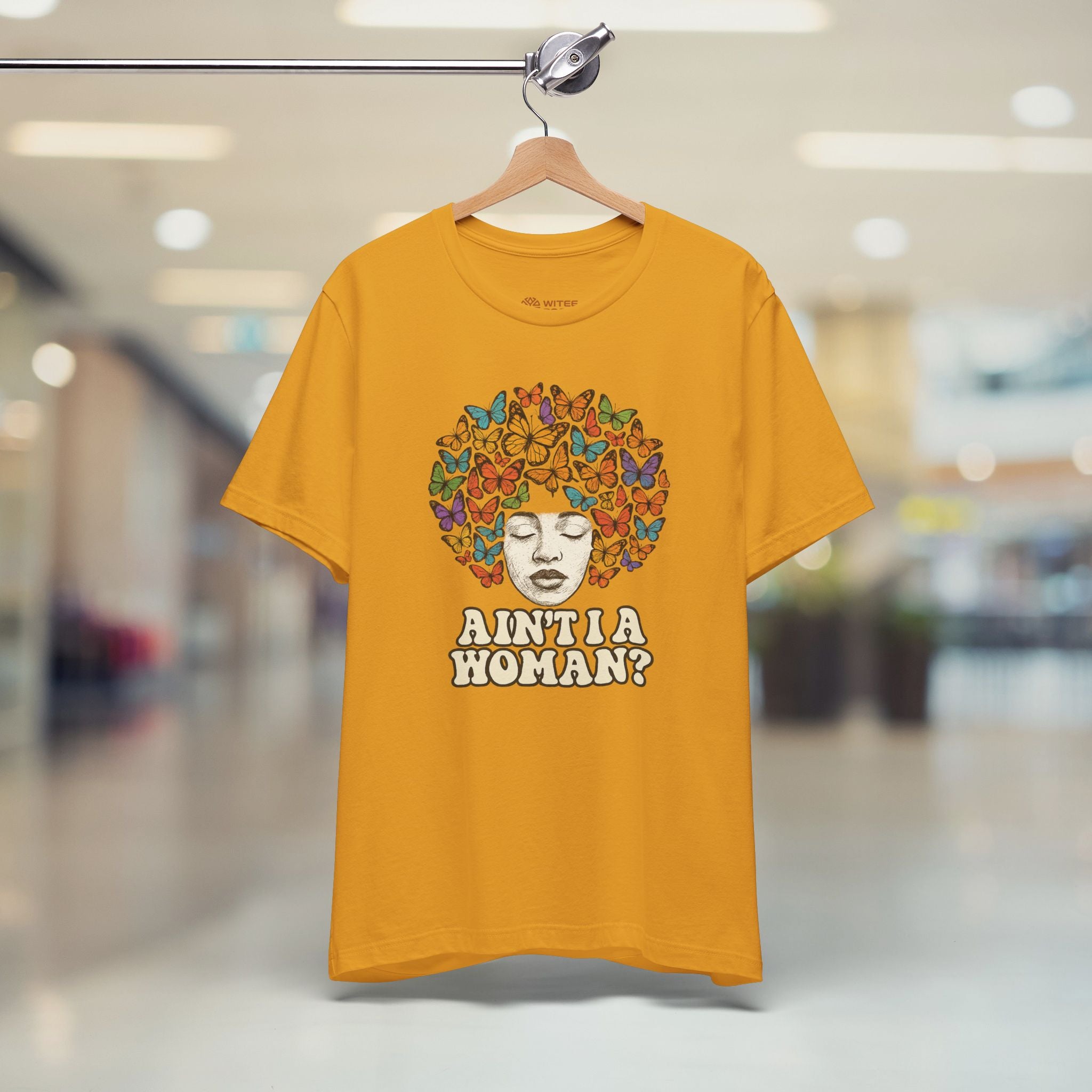 A mustard yellow t-shirt hanging on a wooden hanger from a metal rack, set against a blurred background of a shopping mall corridor. The shirt features a graphic of a face surrounded by colorful butterflies and the text 'AIN'TIA WOMAN?'.