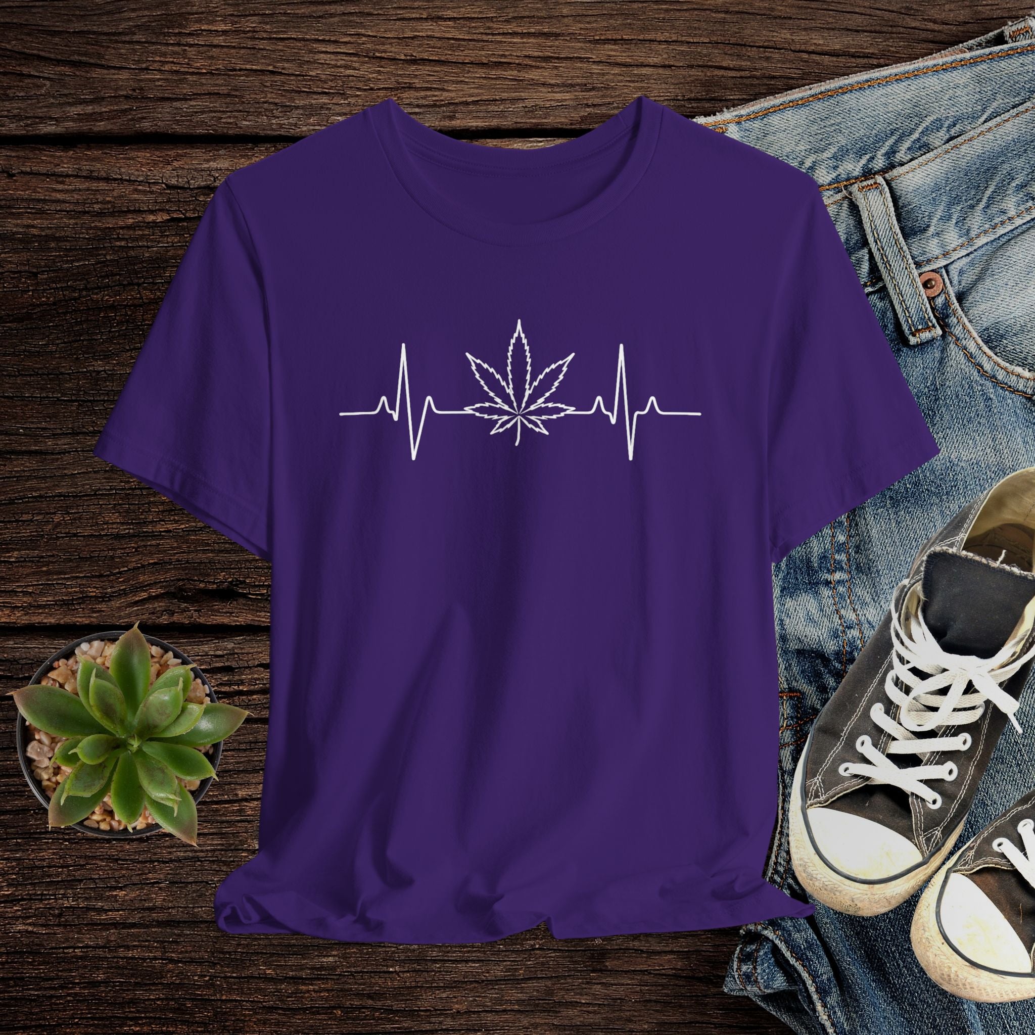 Purple t-shirt with a white cannabis leaf and heartbeat line design on a wooden surface with jeans and sneakers.