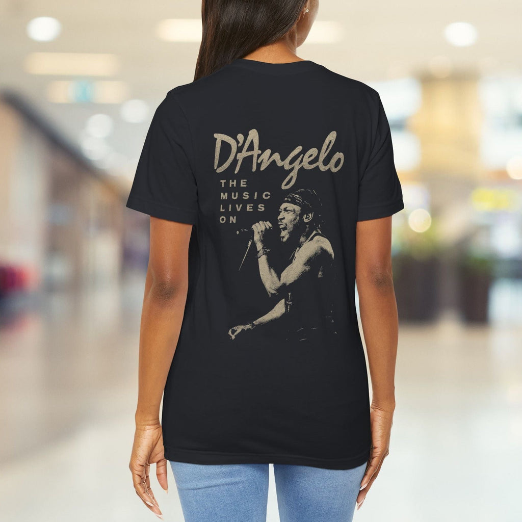 Person wearing a black t-shirt with 'D'Angelo The Music Lives On' design in a blurred indoor setting