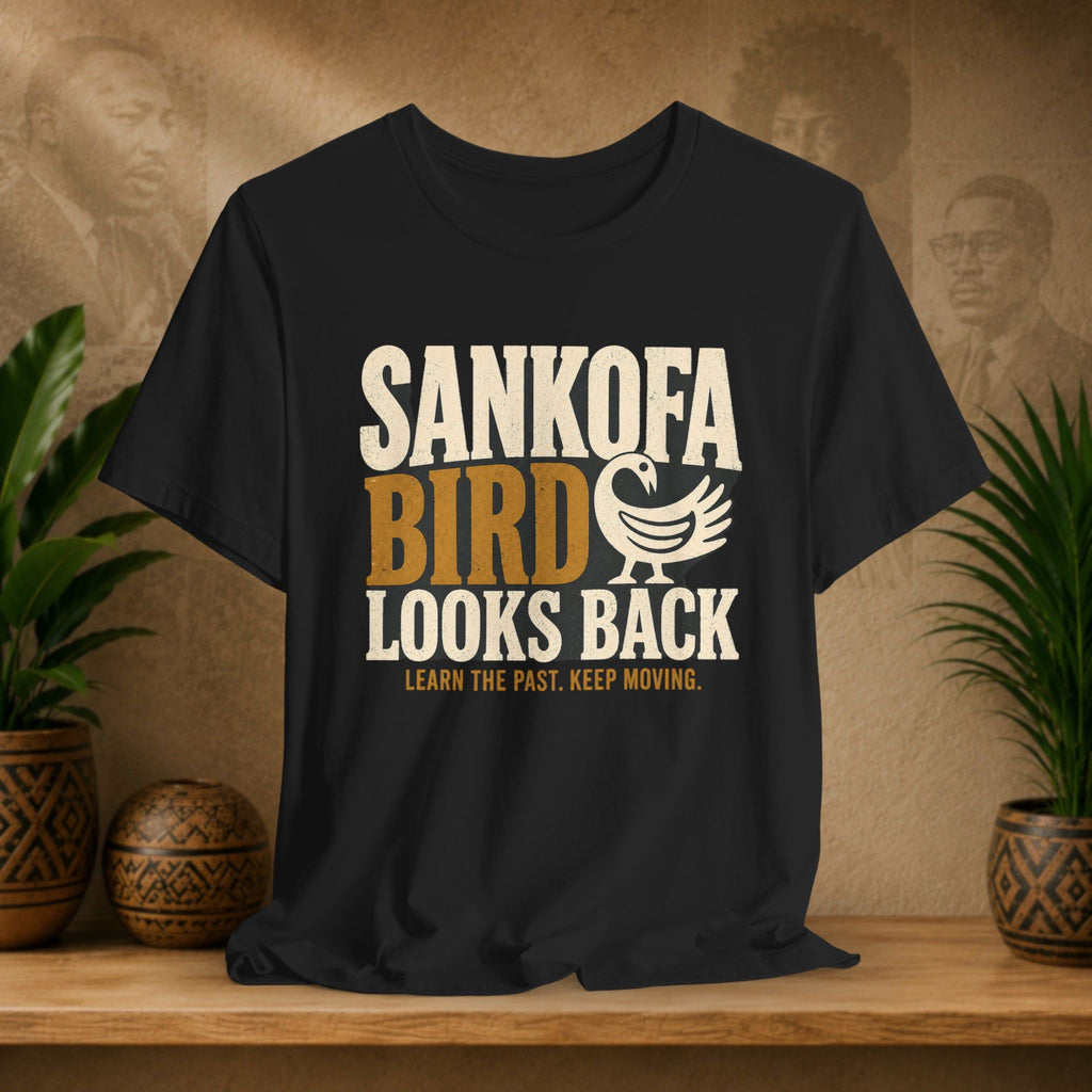 Black t-shirt with 'Sankofa Bird Looks Back' text on a wooden surface with plants and decorative items.