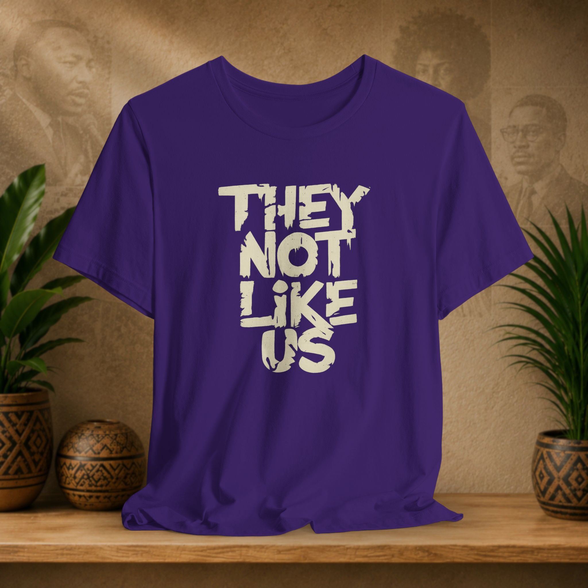 Purple t-shirt with 'THEY NOT LIKE US' text on a wooden surface with plants and a textured wall background