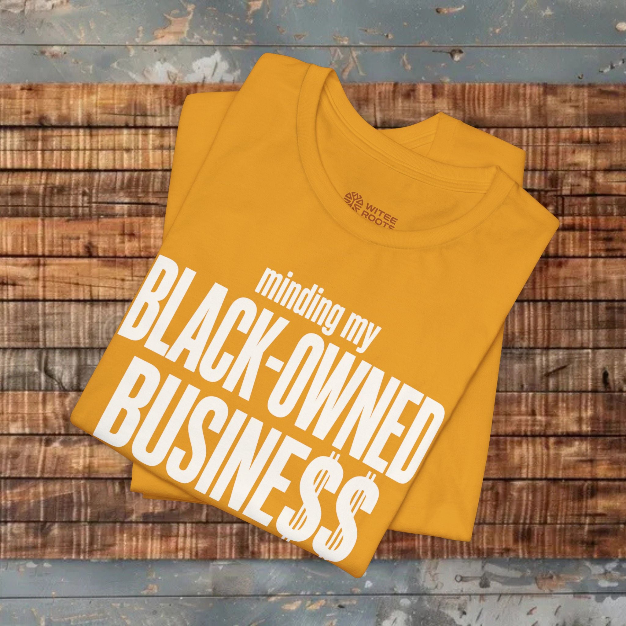 A folded mustard yellow t-shirt with bold white text reading 'minding my BLACK-OWNED BUSINESS$$' is displayed on a rustic wooden plank surface.