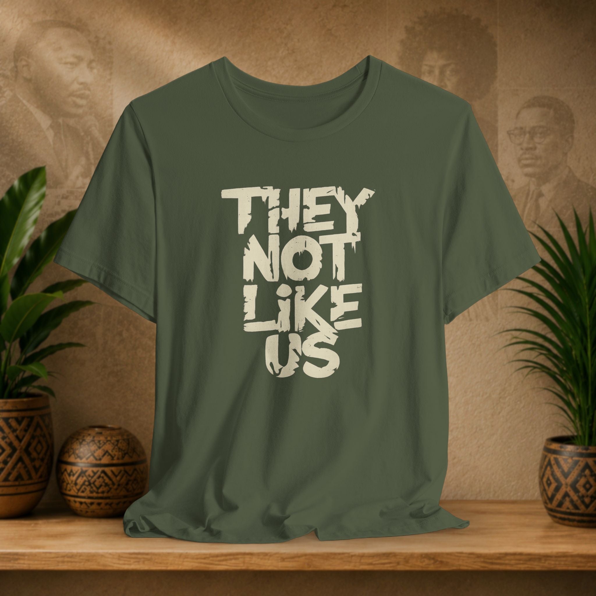 Green t-shirt with 'THEY NOT LIKE US' text on a wooden surface with plants and a textured wall background.