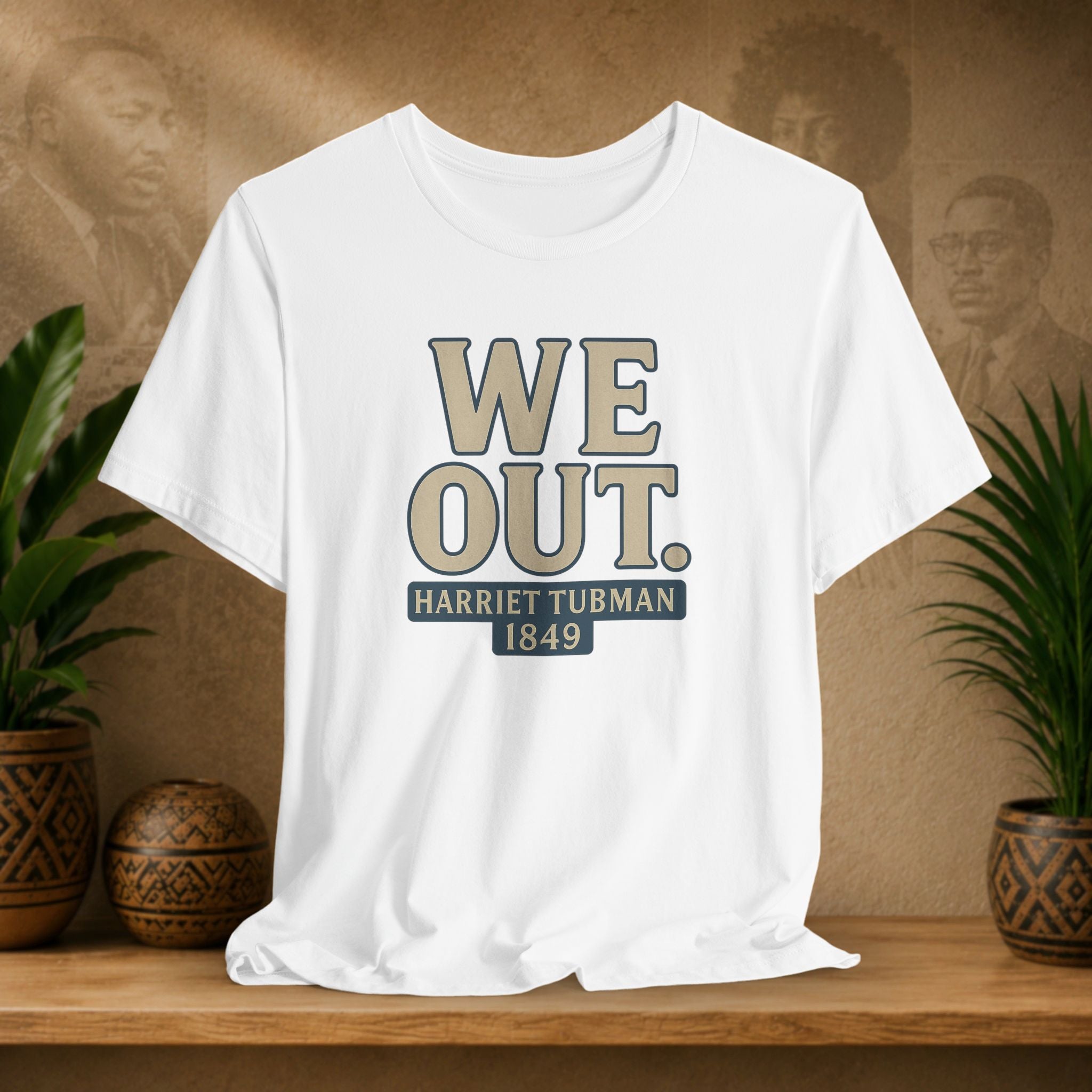 White t-shirt with 'WE OUT. HARRETT TUBMAN 1849' text on a wooden shelf with plants and a blurred background.