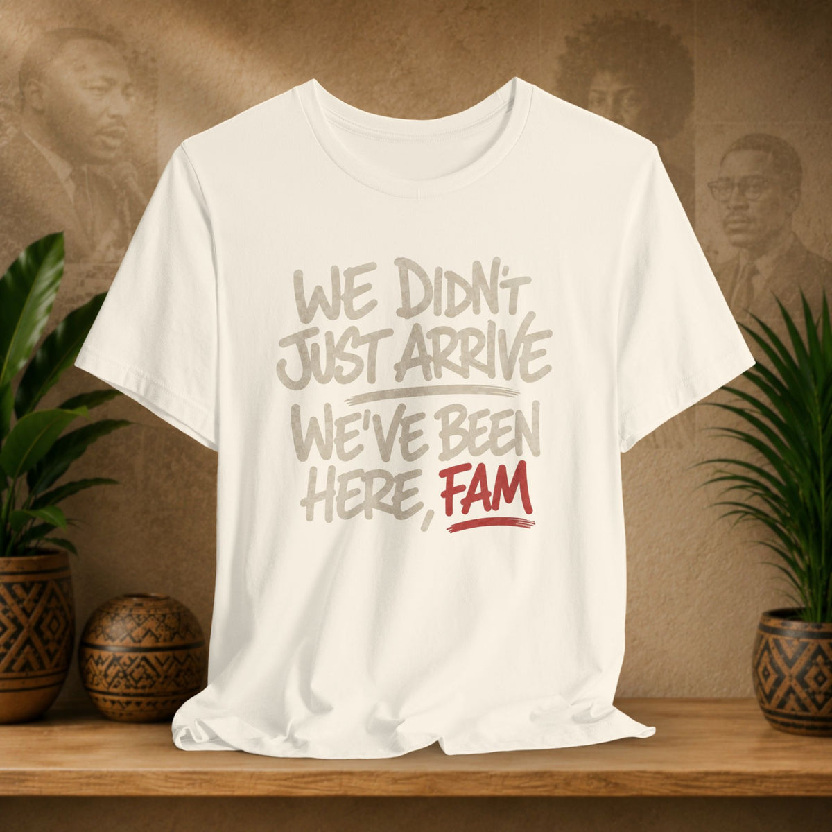 A off-white graphic t-shirt displayed on a wooden shelf, flanked by potted plants and a textured brown background with faded portraits. The shirt features the text 'WE DIDN'T JUST ARRIVE WE'VE BEEN HERE, FAM' in gray and red lettering.
