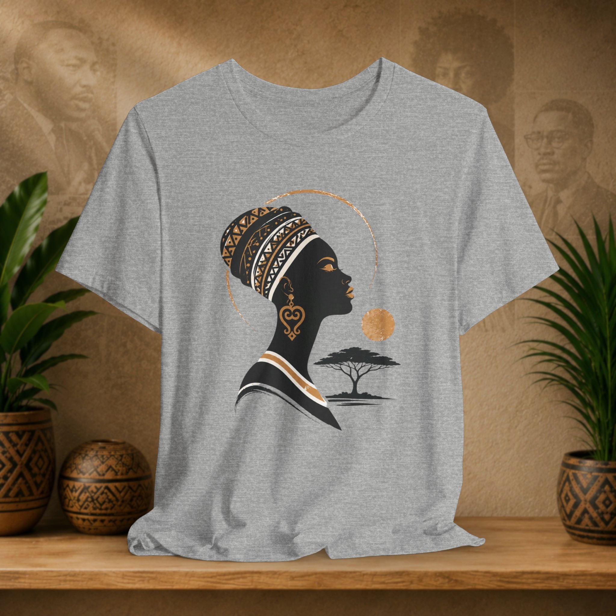 Gray t-shirt with a graphic of a woman's silhouette, sun, and tree on a wooden surface with plants and decorative items.