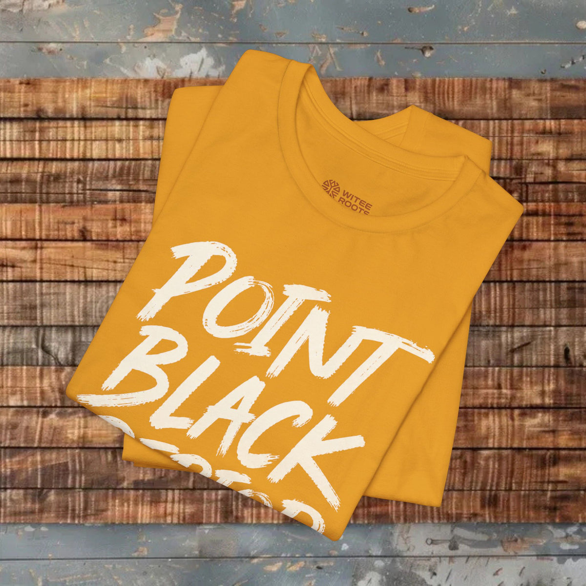 A folded mustard yellow t-shirt with 'POINT BLACK GOOD' printed in white brush lettering, displayed on a rustic wooden plank surface.
