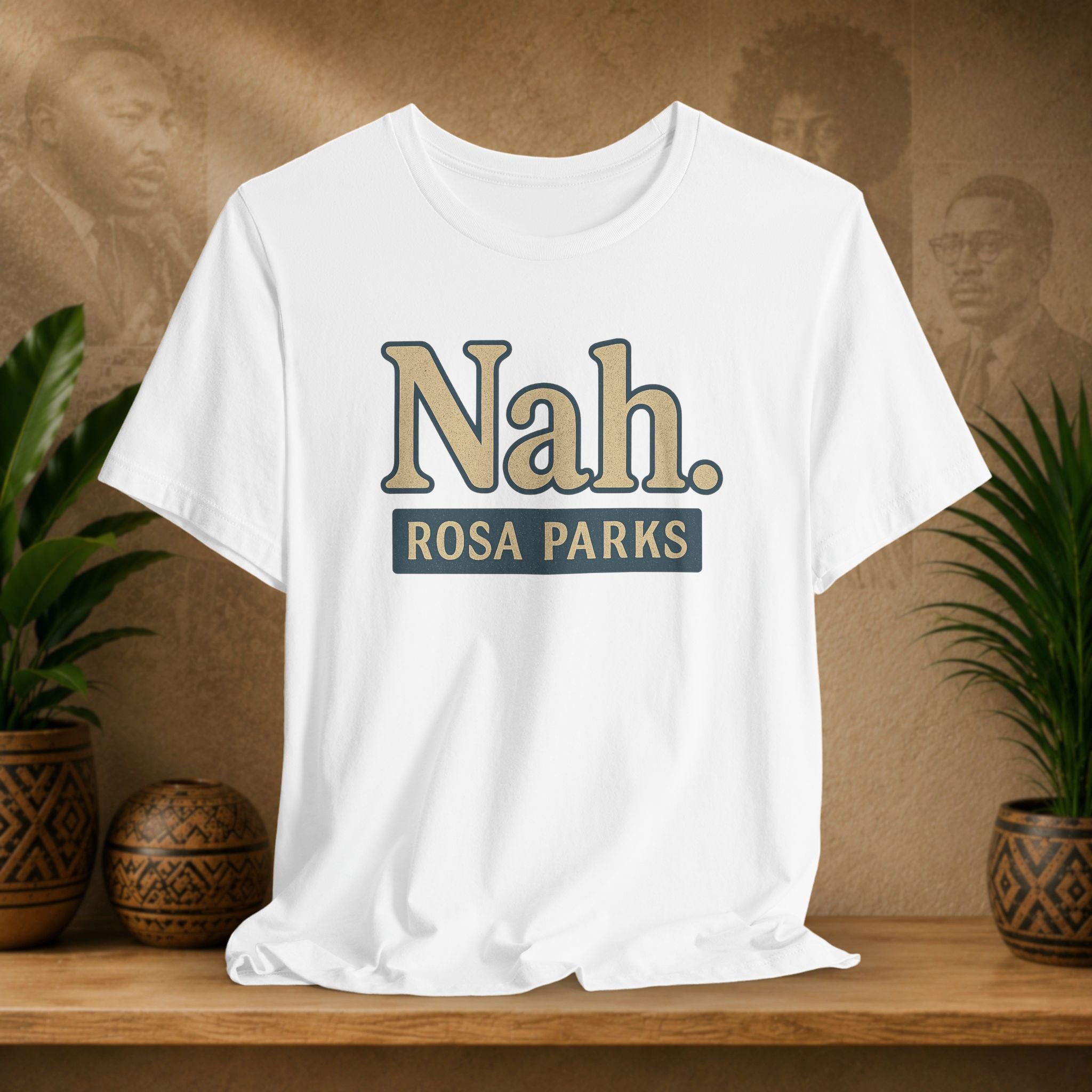 White t-shirt with 'Nah. ROSA PARKS' text on a wooden surface with plants and a textured wall background.