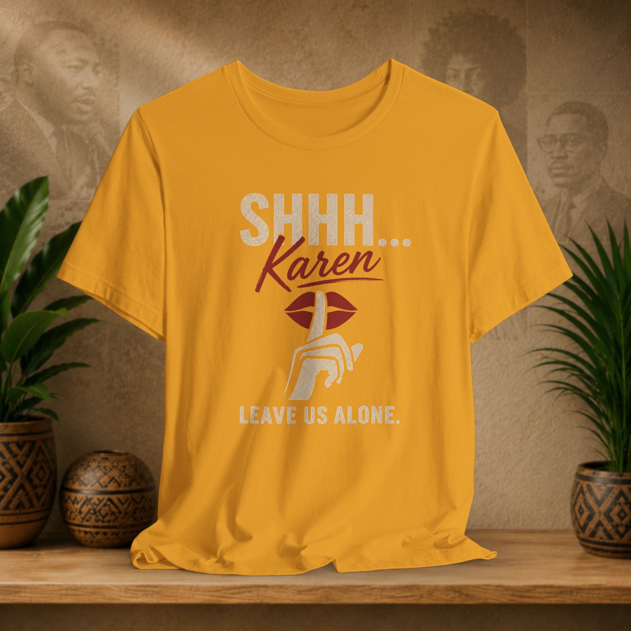A mustard yellow graphic t-shirt displayed on a wooden shelf with potted plants and a textured background. The shirt features a graphic print that reads 'SHHH.
