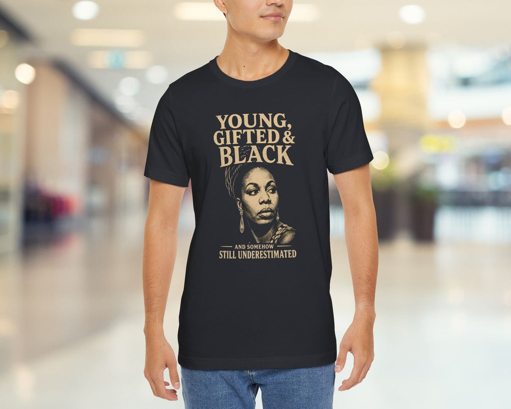 A man wearing a black graphic t-shirt with a gold and white print that reads 'YOUNG, GIFTED & BLACK' and 'JEN NASHOR IS STILL UNDERESTIMATED', standing in a blurred indoor hallway.