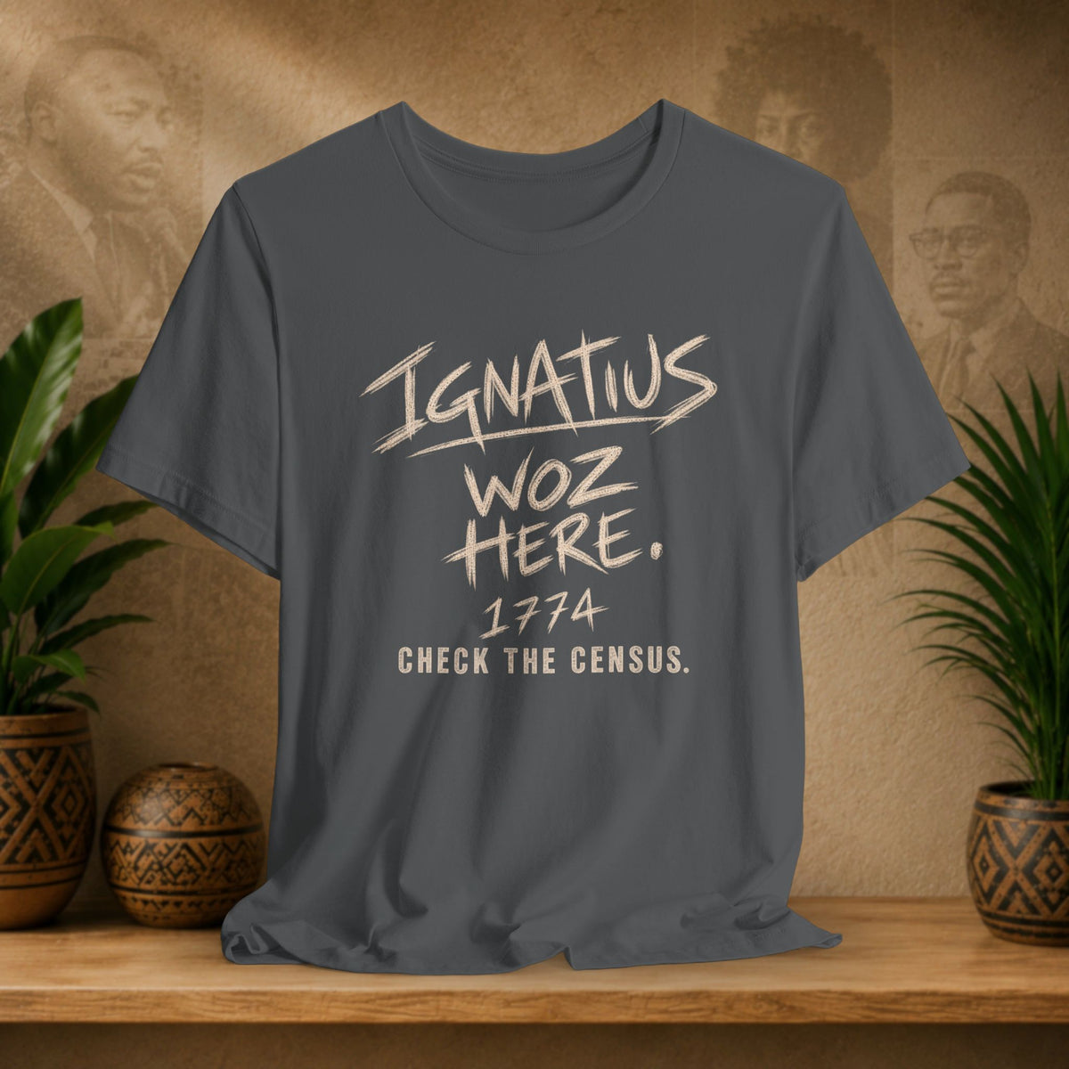 A dark gray graphic t-shirt displayed on a wooden shelf, flanked by potted plants and a textured background with sepia portraits. The shirt features the text 'IGNATIUS WOZ HERE.