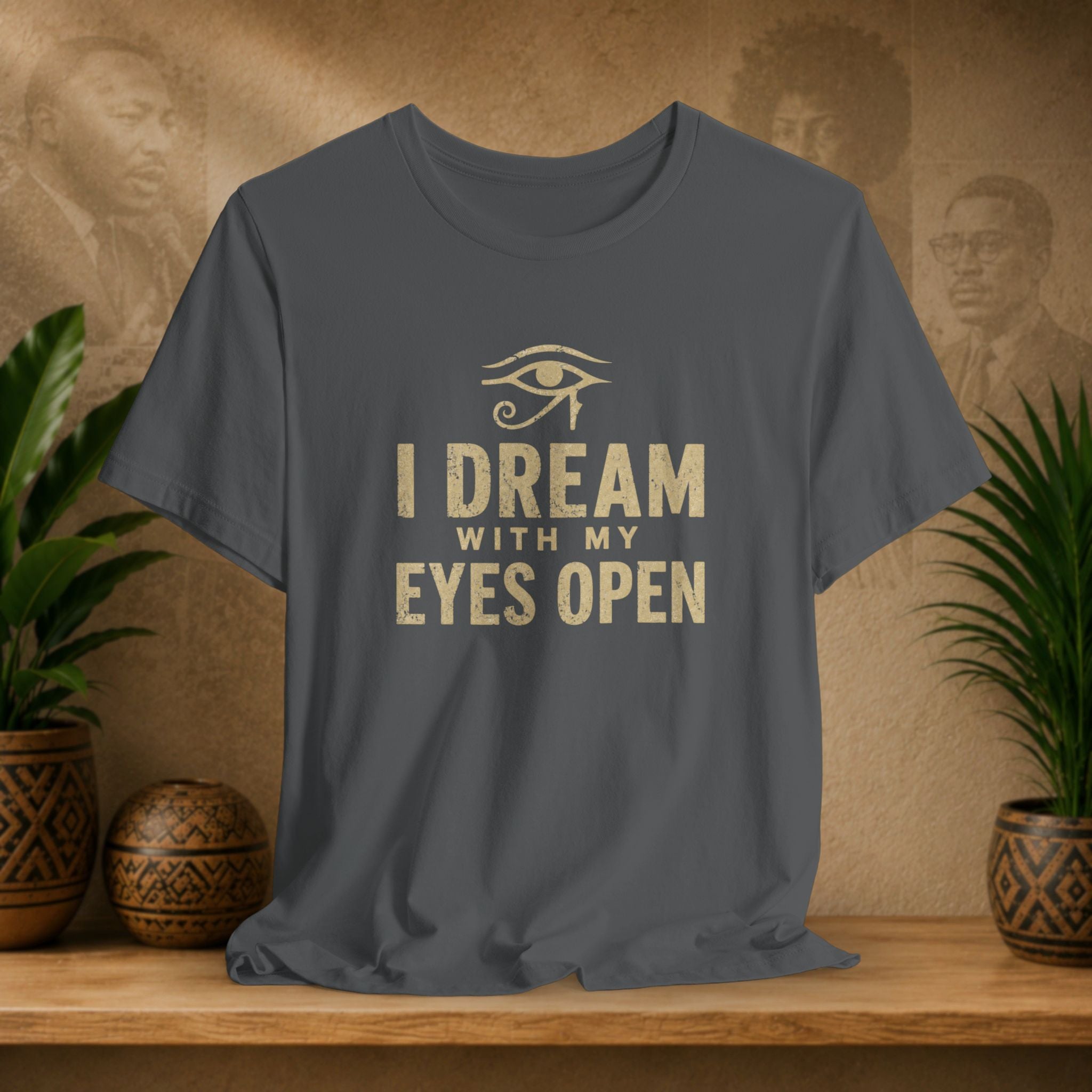 Gray t-shirt with 'I Dream with My Eyes Open' text and eye symbol on a wooden surface with plants and decorative items.