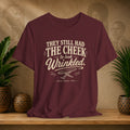 Maroon t-shirt with white text on a wooden surface with plants and a textured wall background