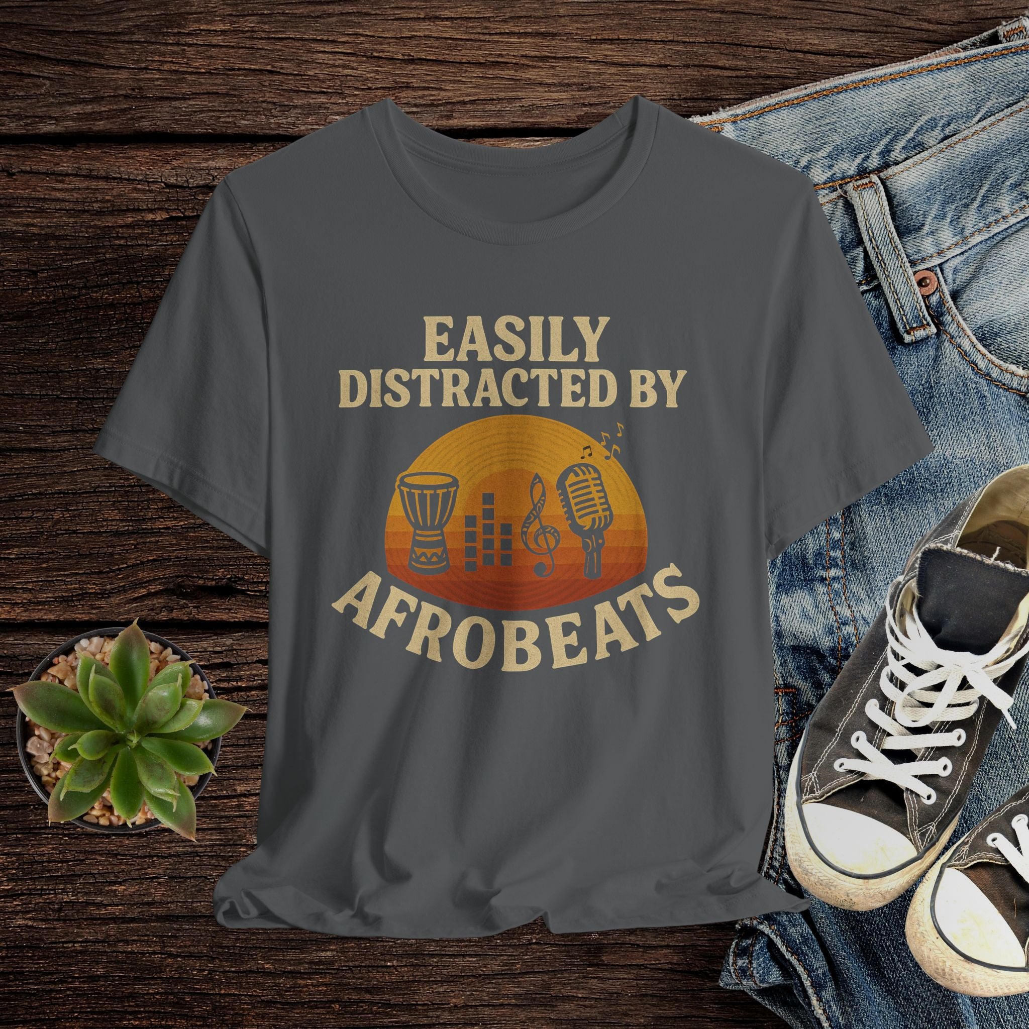 Gray t-shirt with 'Easily Distracted by AfroBeats' design on a wooden surface with jeans and sneakers.