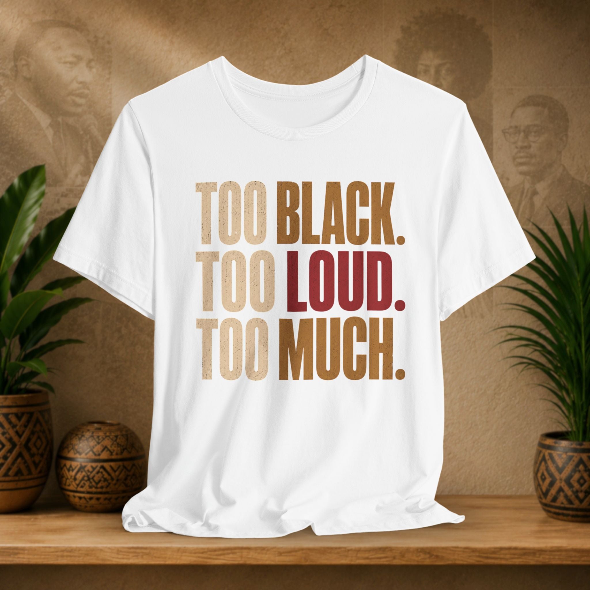 A white short-sleeve graphic T-shirt displayed on a wooden surface, featuring the text 'TOO BLACK. TOO LOUD.