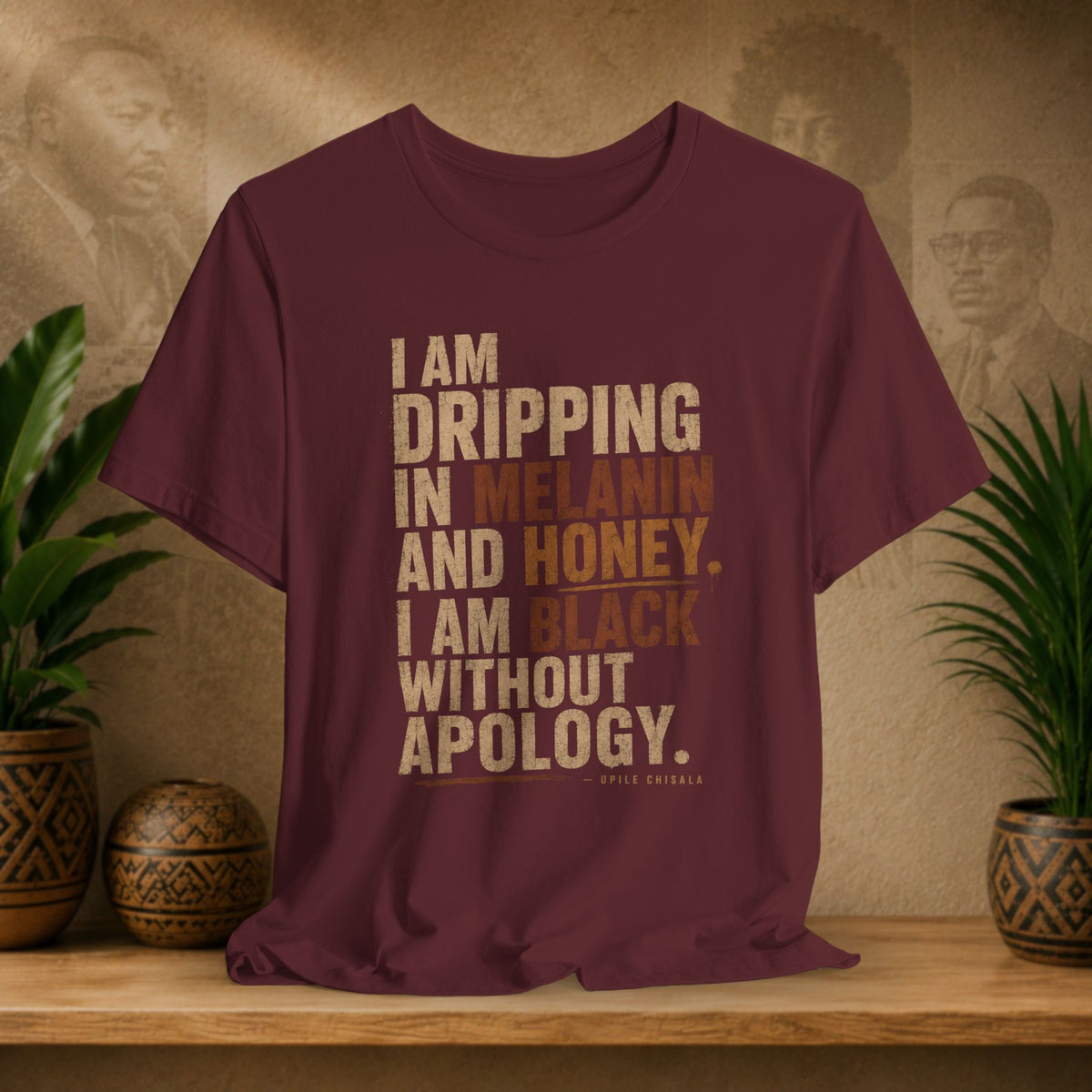 A maroon short-sleeve graphic T-shirt displayed on a wooden shelf, featuring a stacked text design in tan, brown, and cream tones. The text reads: 'I AM DRIPPING IN MELANIN AND HONEY.