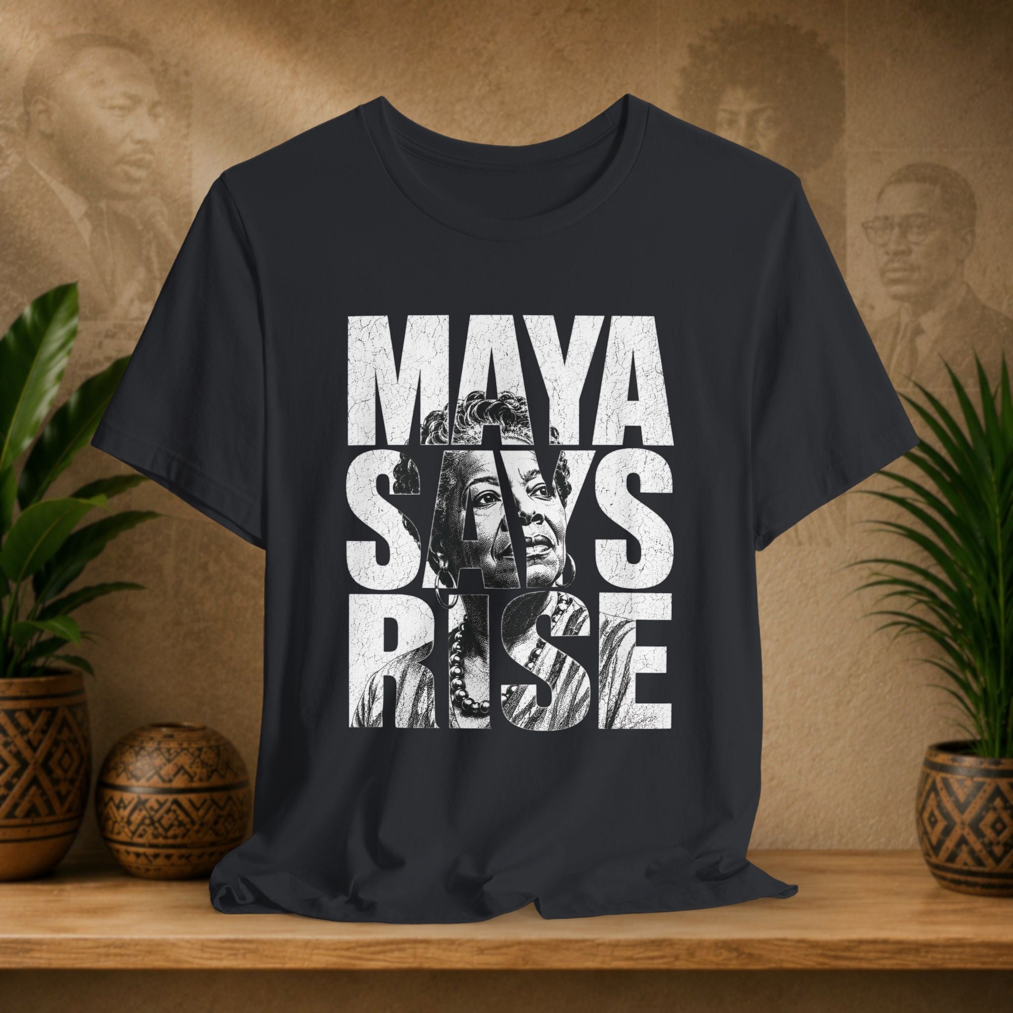 Black t-shirt with 'MAYA SAYS RISE' text and graphic on a wooden surface with plants and decorative items.