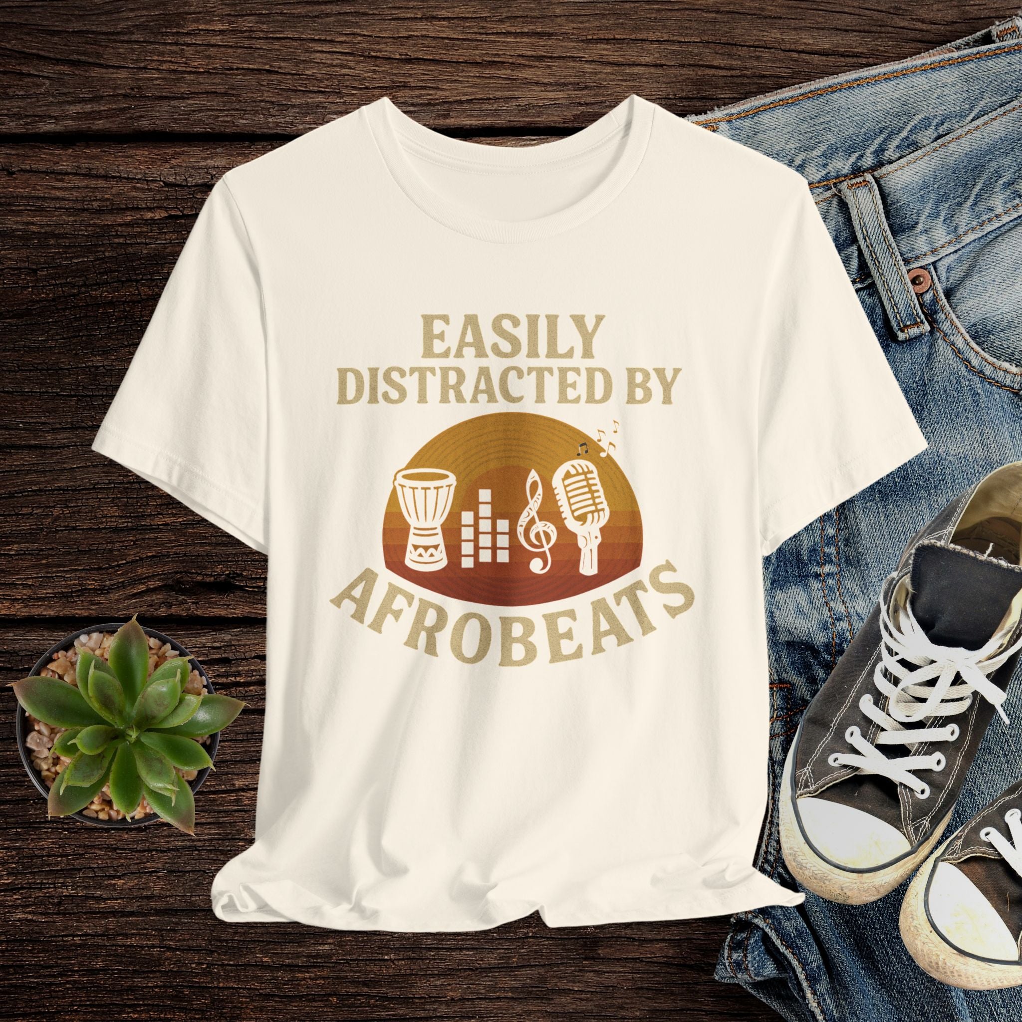 Beige t-shirt with 'Easily Distracted by AfroBeats' design on a wooden surface with jeans and sneakers.