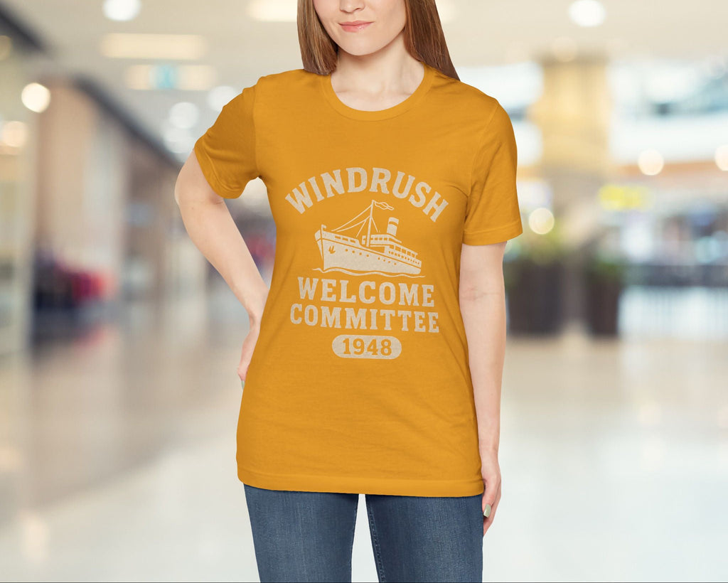 Woman wearing a mustard yellow t-shirt with 'Windrush Welcome Committee 1948' text and graphics in a blurred indoor setting.
