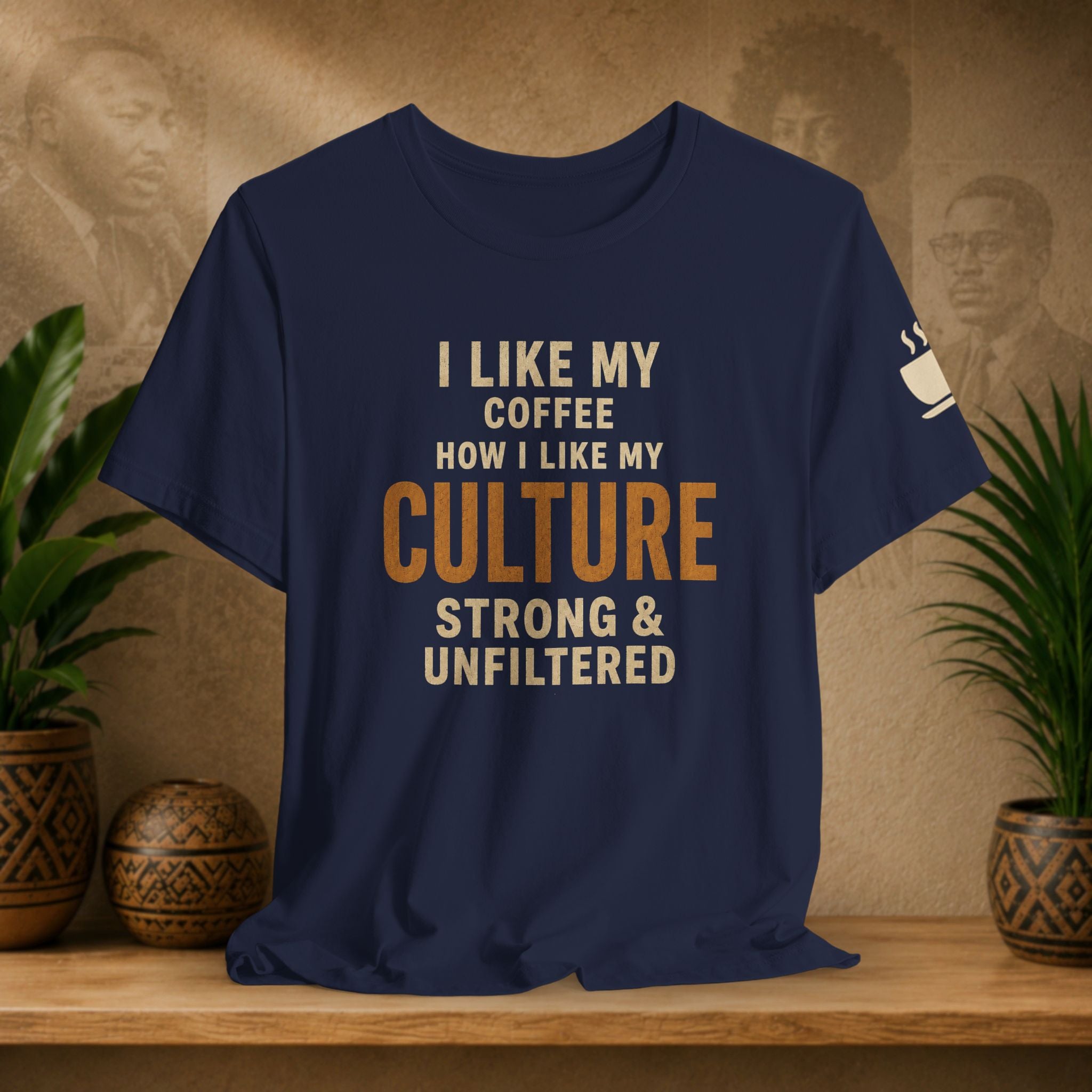 A navy blue graphic t-shirt displayed on a wooden shelf with potted plants and a textured beige background. The shirt features the text 'I LIKE MY COFFEE HOW I LIKE MY CULTURE STRONG & UNFILTERED' in cream and orange lettering, with a small white coffee cup graphic on the left sleeve.
