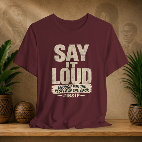Maroon t-shirt with text 'Say it LOUD Enough for the people in the back #IB&IP' on a wooden surface with a vintage-style background.