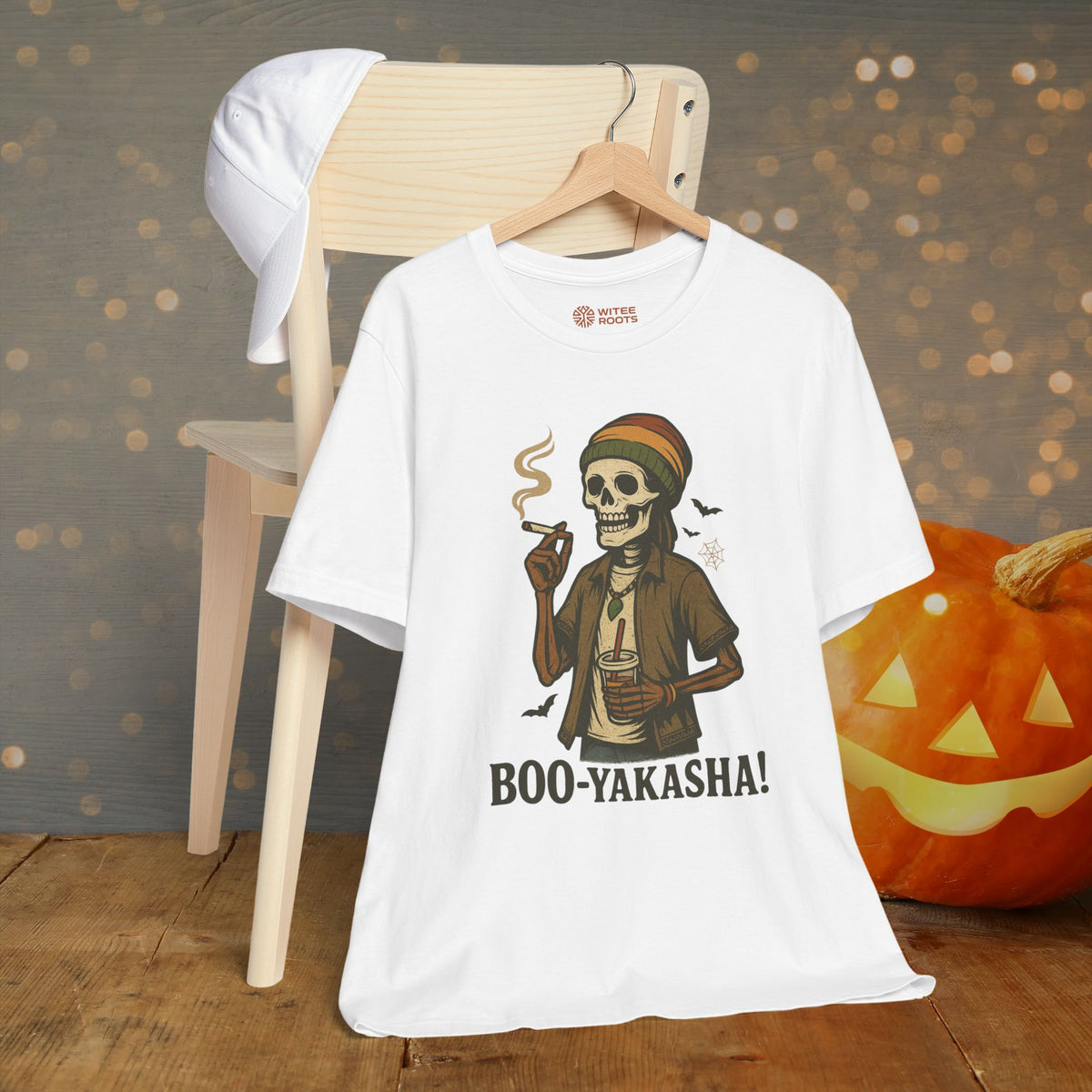 A white graphic T-shirt hanging on a wooden hanger, featuring a cartoon skeleton with a Rasta hat smoking and holding a cup, with the text 'BOO-YAKASHA!' below. The shirt is styled in a Halloween-themed setting with a wooden chair, a white cap, and a carved pumpkin in the background.