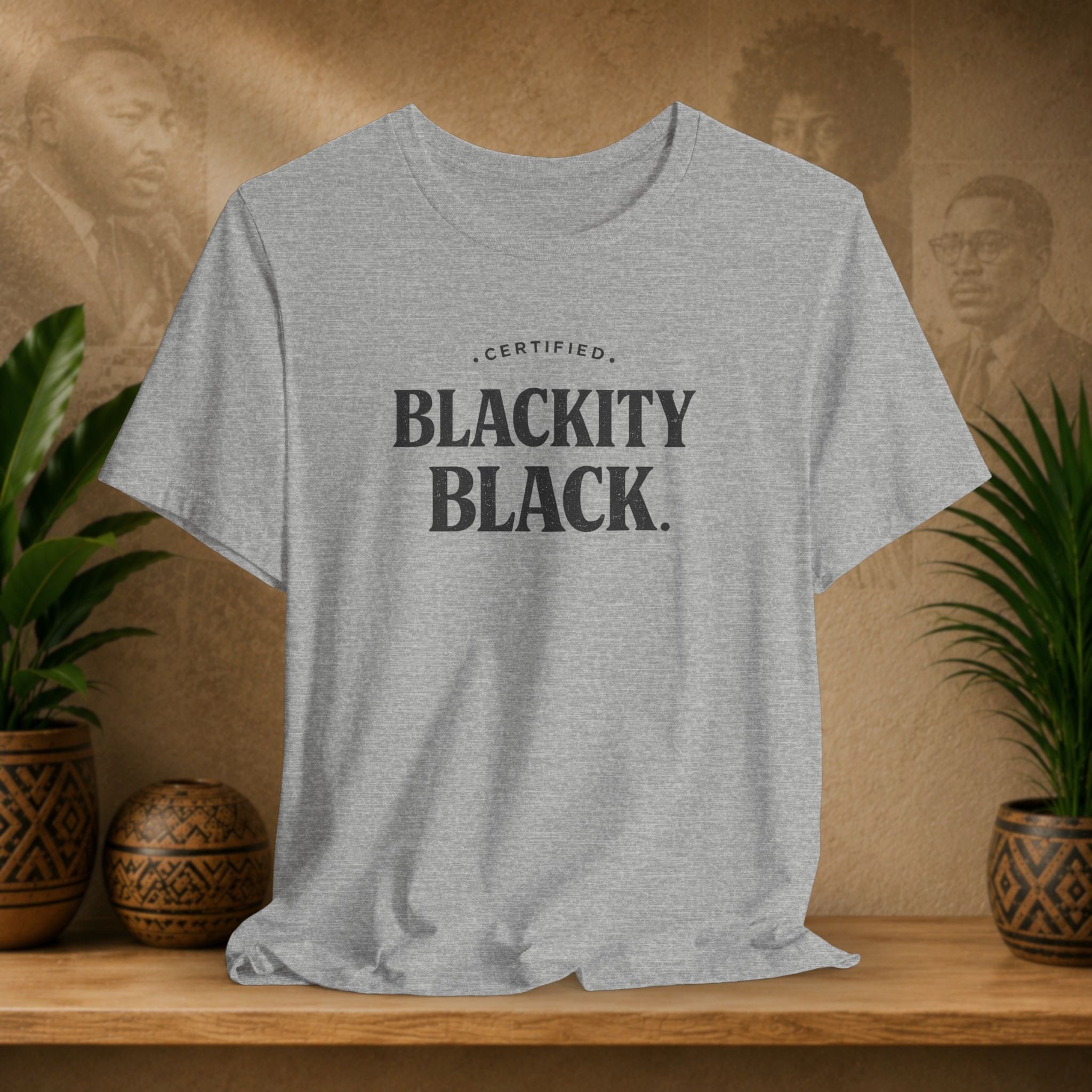 Gray t-shirt with 'CERTIFIED BLACKITY BLACK.' text on a wooden surface with plants and a decorative wall in the background.