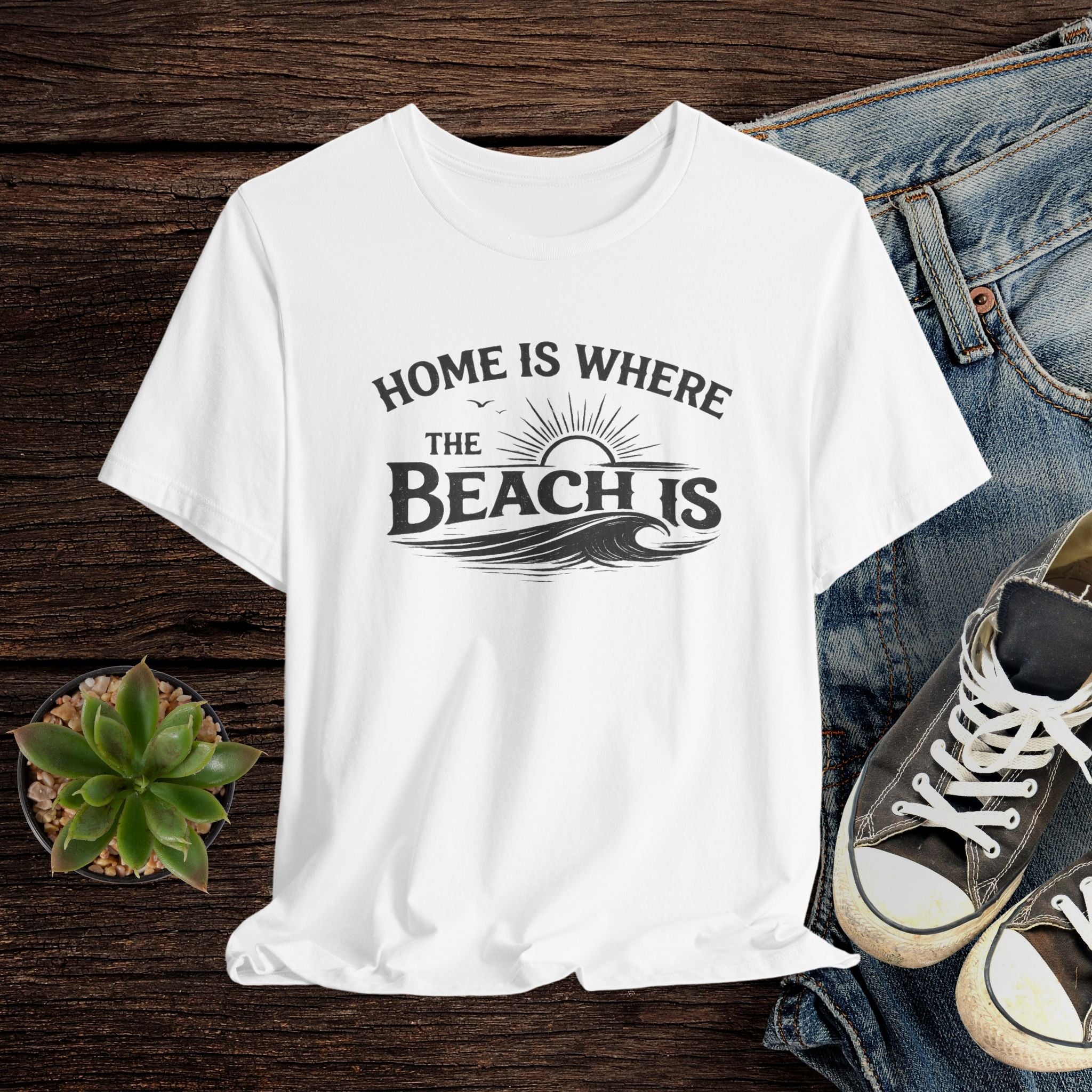 White t-shirt with 'Home is where the beach is' text on a wooden surface with jeans and sneakers.