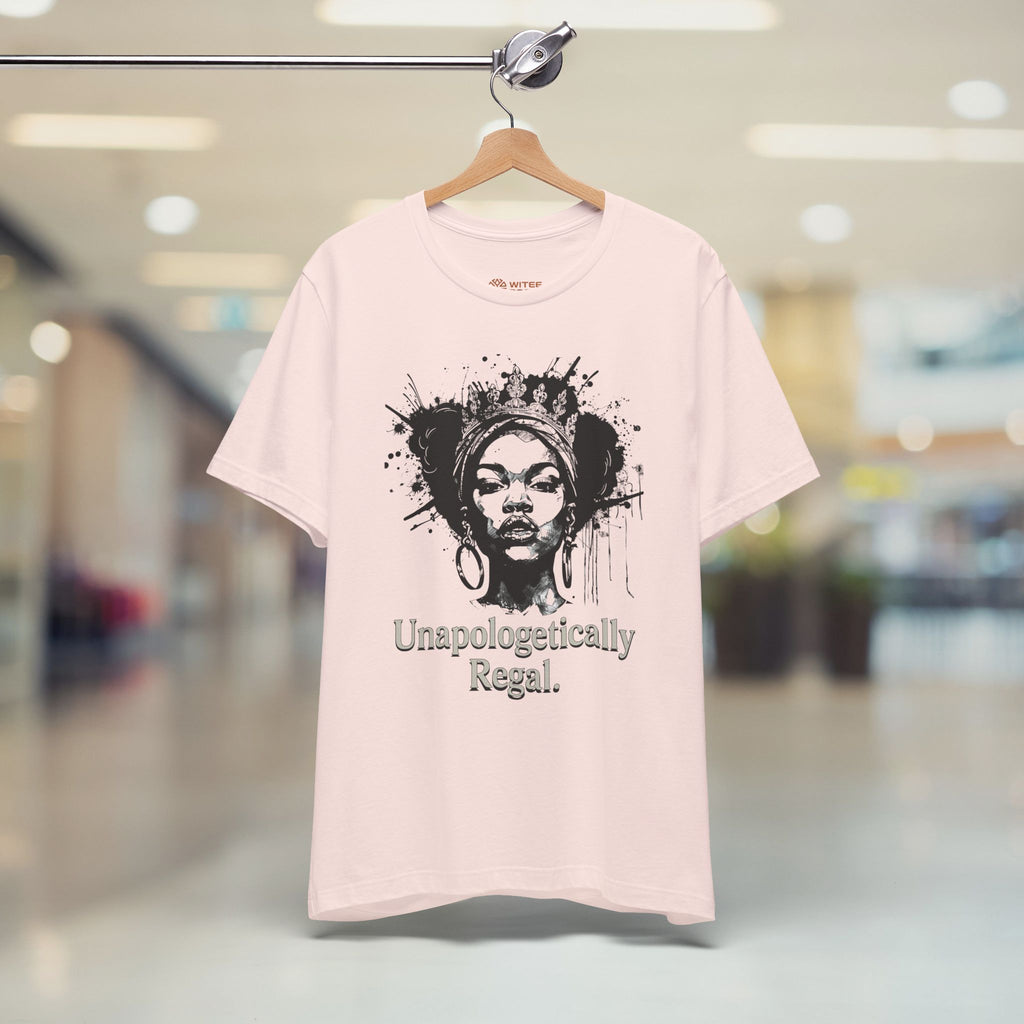 A light pink t-shirt hanging on a wooden hanger from a metal rod, set against a blurred indoor background. The shirt features a black graphic of a woman's face with a headwrap and paint splatters, with the text 'Unapologetically Regal.