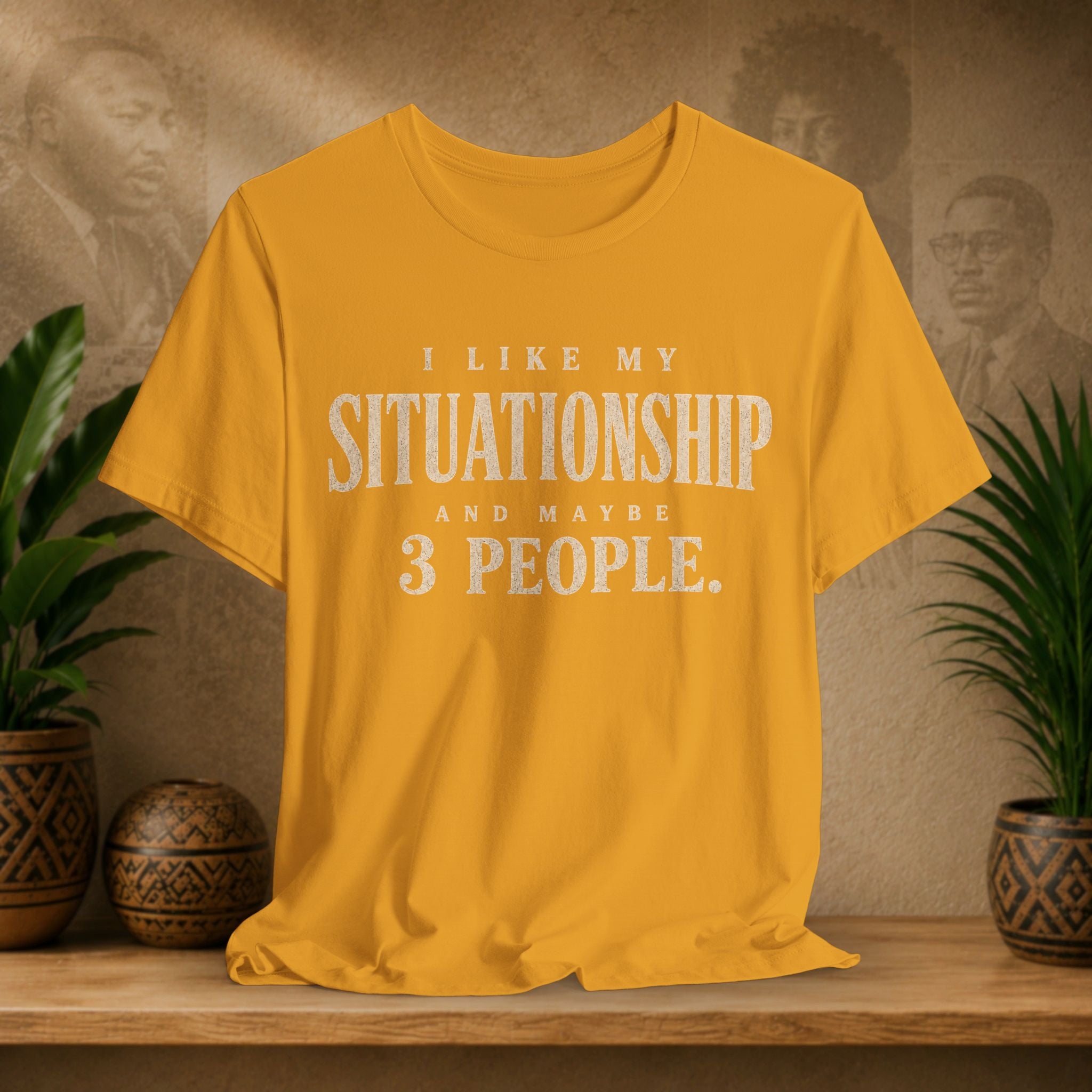 A mustard yellow short-sleeve t-shirt with distressed white text reading 'I LIKE MY SITUATIONSHIP AND MAYBE 3 PEOPLE.' displayed on a wooden shelf with potted plants and a textured background.