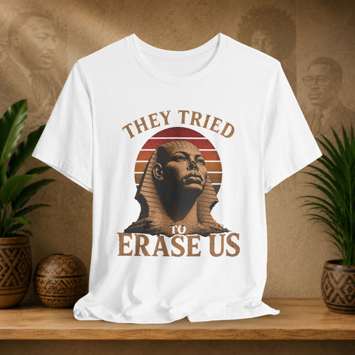 white t-shirt with a graphic of a sphinx and text 'They tried to erase us' on a wooden surface with plants and a textured wall in the background.
