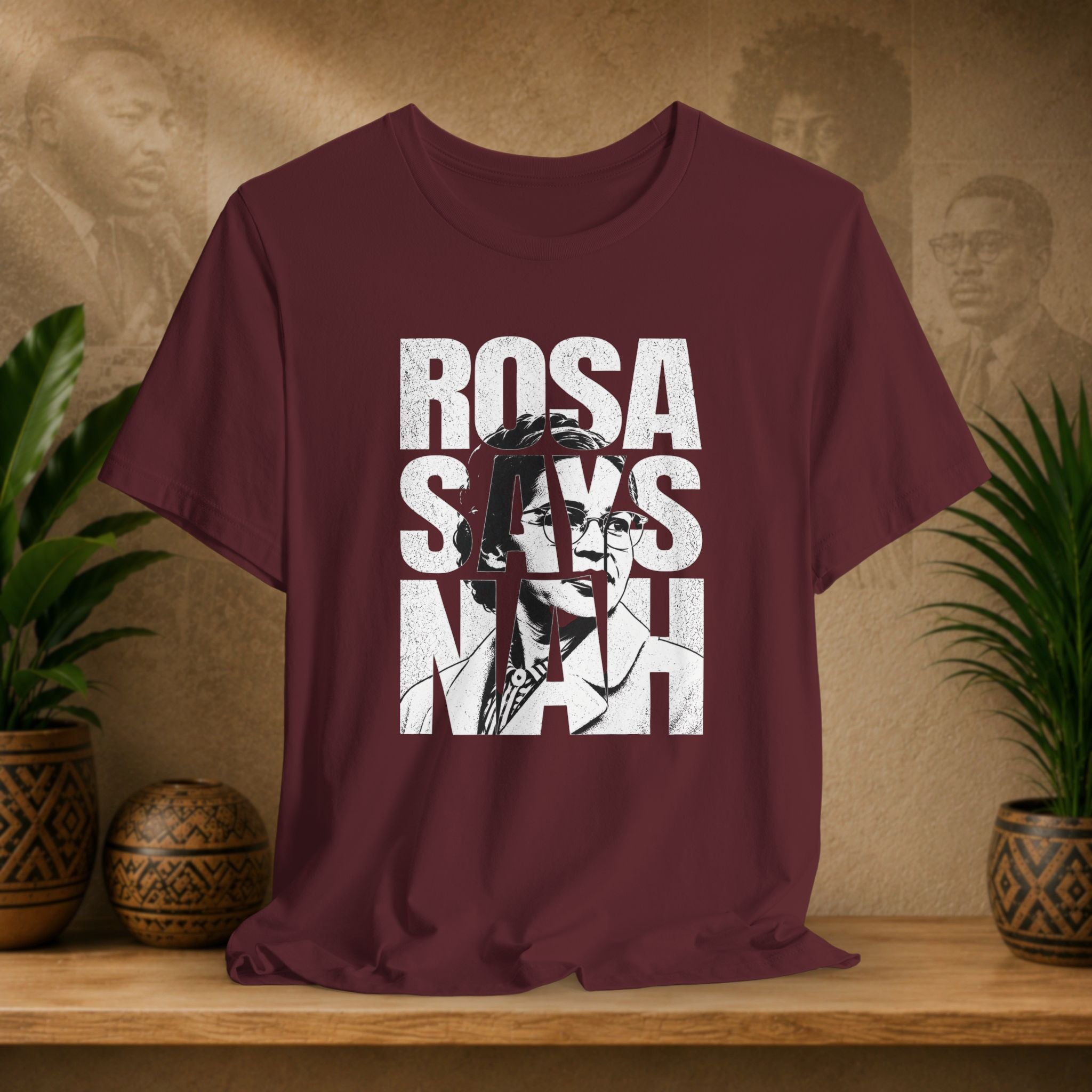 Maroon t-shirt with 'Rosa Parks' graphic design on a wooden surface with plants and a textured wall in the background.