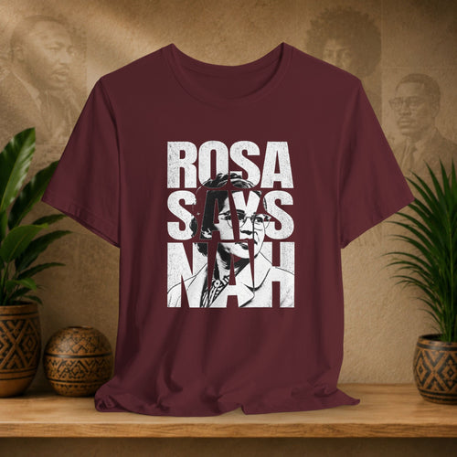 Maroon t-shirt with 'Rosa Parks' graphic design on a wooden surface with plants and a textured wall in the background.