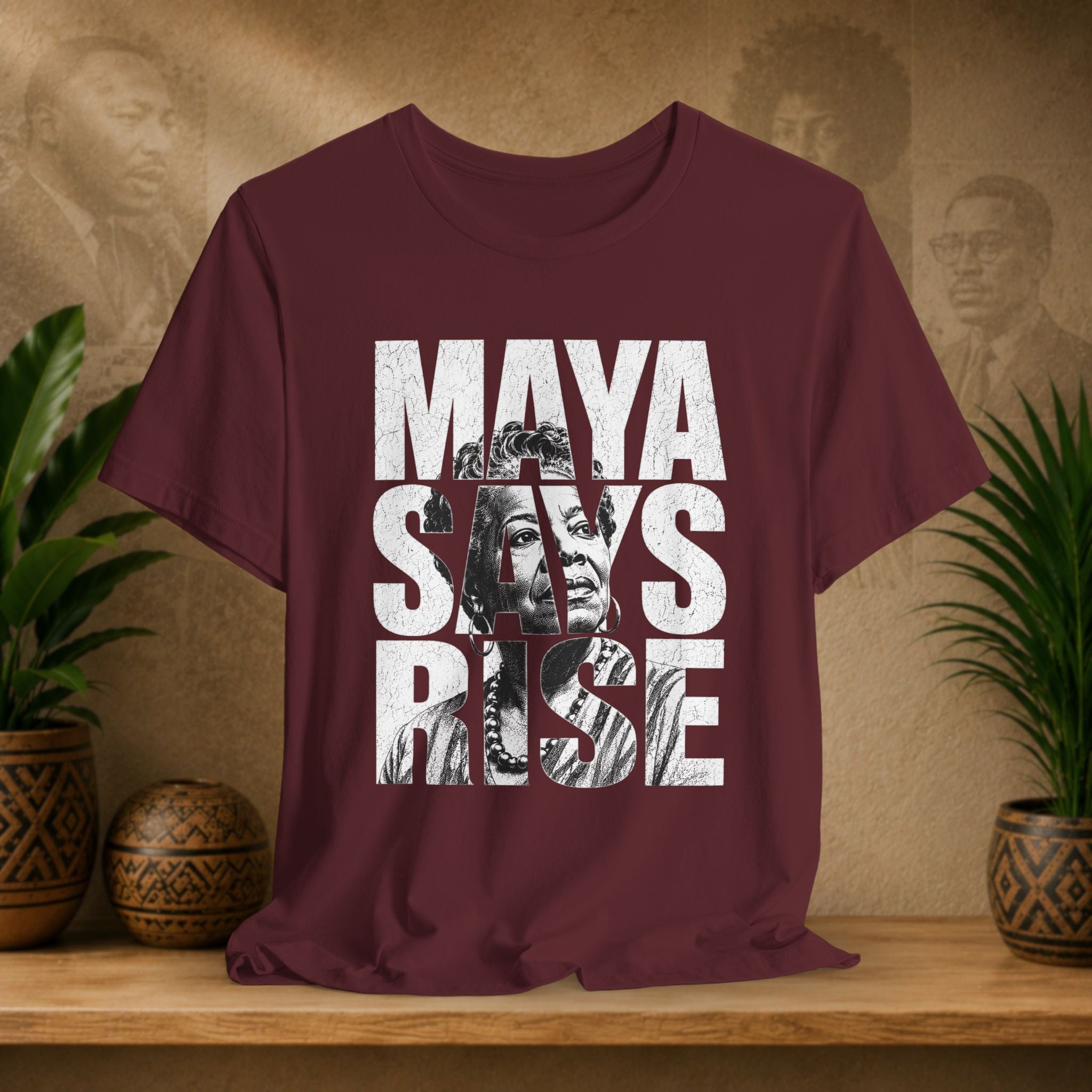 Maroon t-shirt with 'MAYA SAYS RISE' text and images on a wooden surface with plants and decorative items.