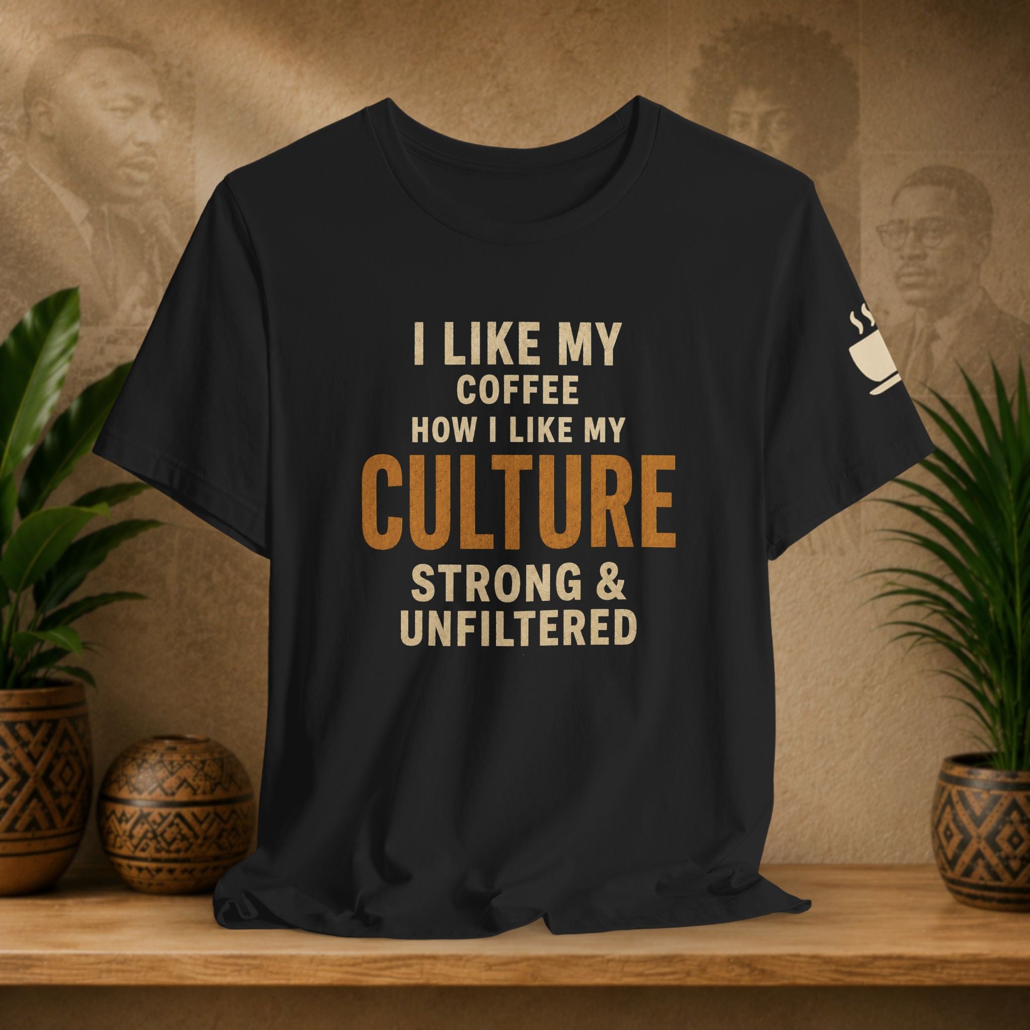 A black graphic t-shirt displayed on a wooden shelf, flanked by potted green plants. The shirt features the text 'I LIKE MY COFFEE HOW I LIKE MY CULTURE STRONG & UNFILTERED' in cream and orange lettering, with a small white coffee cup graphic on the left sleeve.