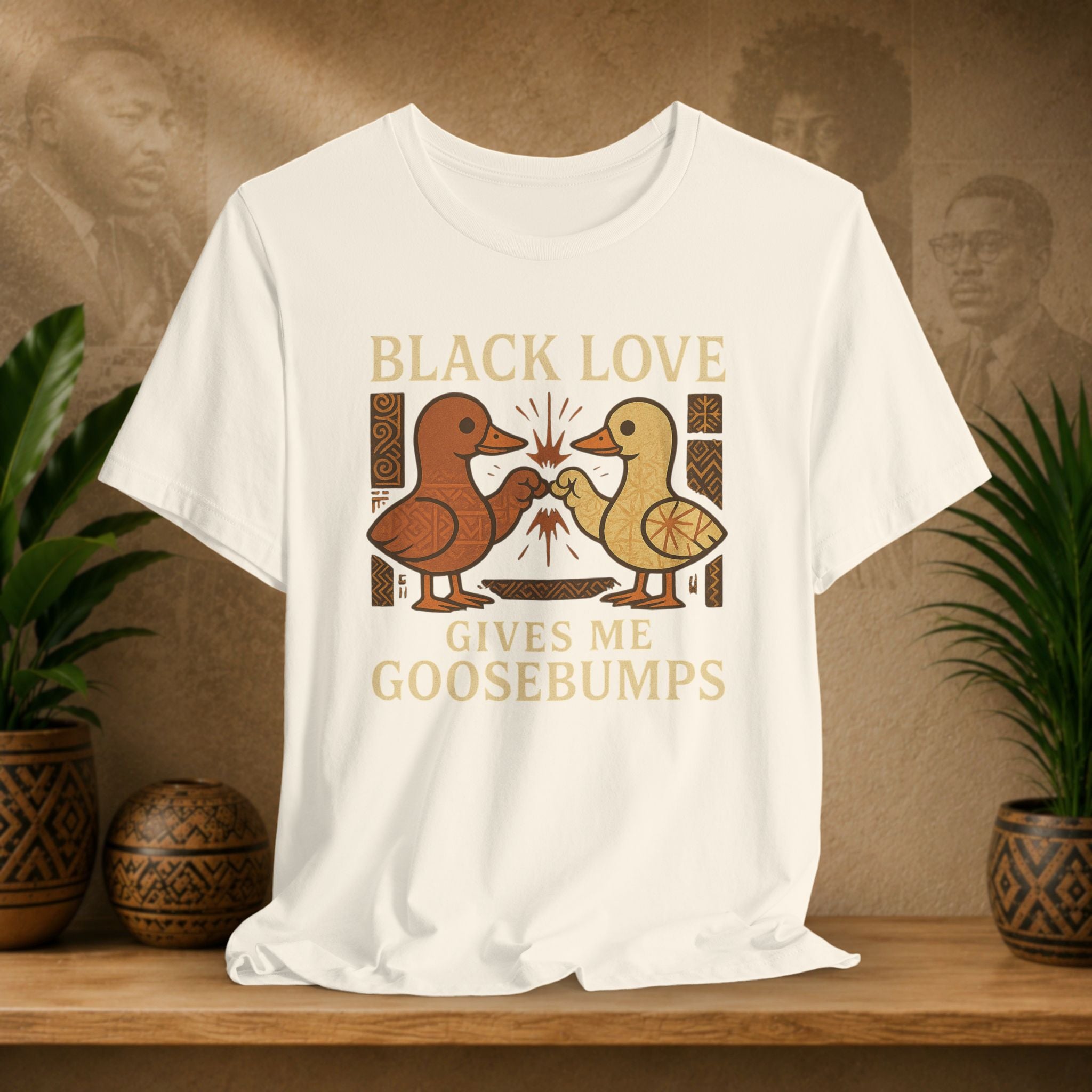 Beige t-shirt with 'Black Love Gives Me Goosebumps' graphic on a wooden surface with plants and a textured wall background.