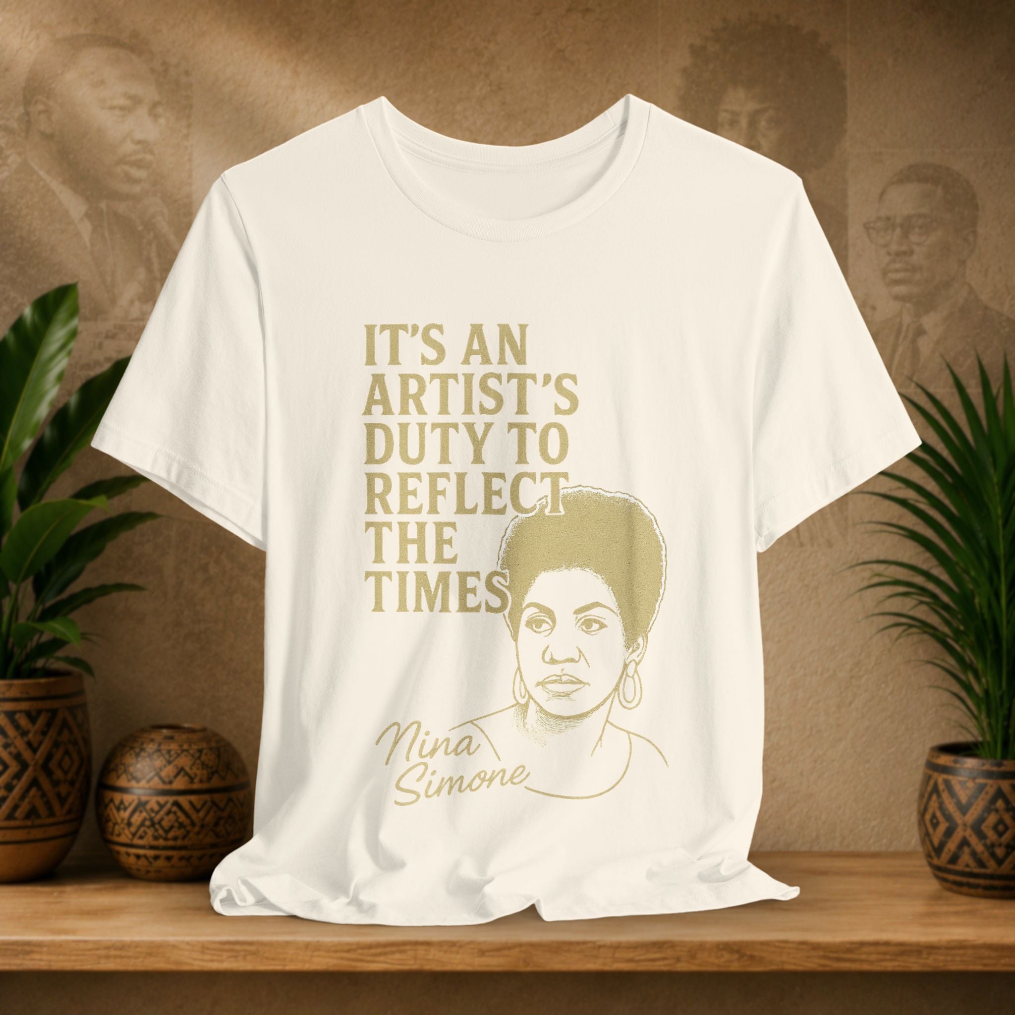 Beige t-shirt with gold text and Nina Simone's portrait on a wooden surface with plants and a blurred background.