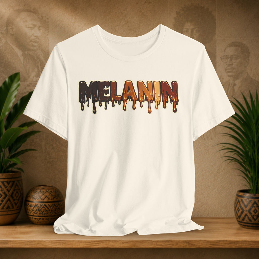 A beige short-sleeve crewneck t-shirt with a dripping 'MELANIN' graphic in dark brown, orange, and black, displayed on a wooden shelf with potted plants and a textured background.