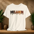 A beige short-sleeve crewneck t-shirt with a dripping 'MELANIN' graphic in dark brown, orange, and black, displayed on a wooden shelf with potted plants and a textured background.