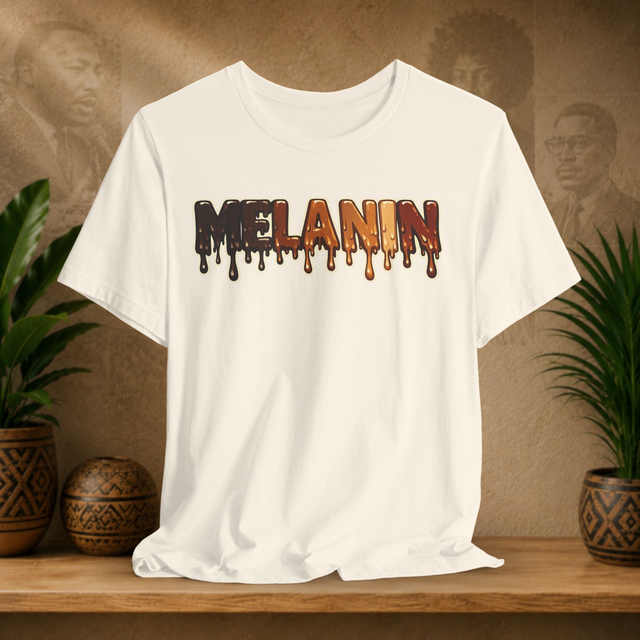 A beige short-sleeve crewneck t-shirt with a dripping 'MELANIN' graphic in dark brown, orange, and black, displayed on a wooden shelf with potted plants and a textured background.