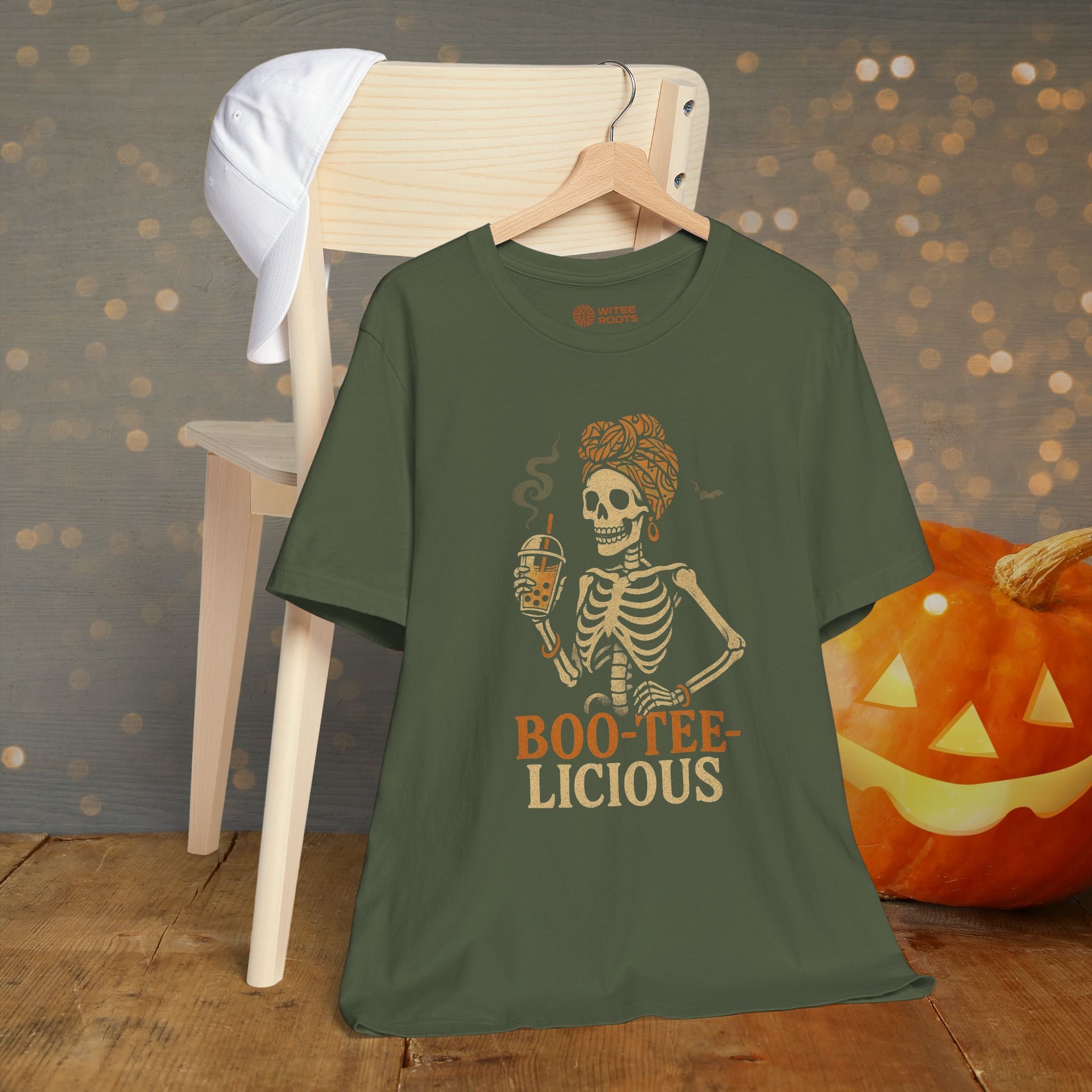 An olive green graphic t-shirt hanging on a wooden hanger, styled in a Halloween-themed setting with a wooden chair, a white baseball cap, and a carved jack-o'-lantern pumpkin against a bokeh background.