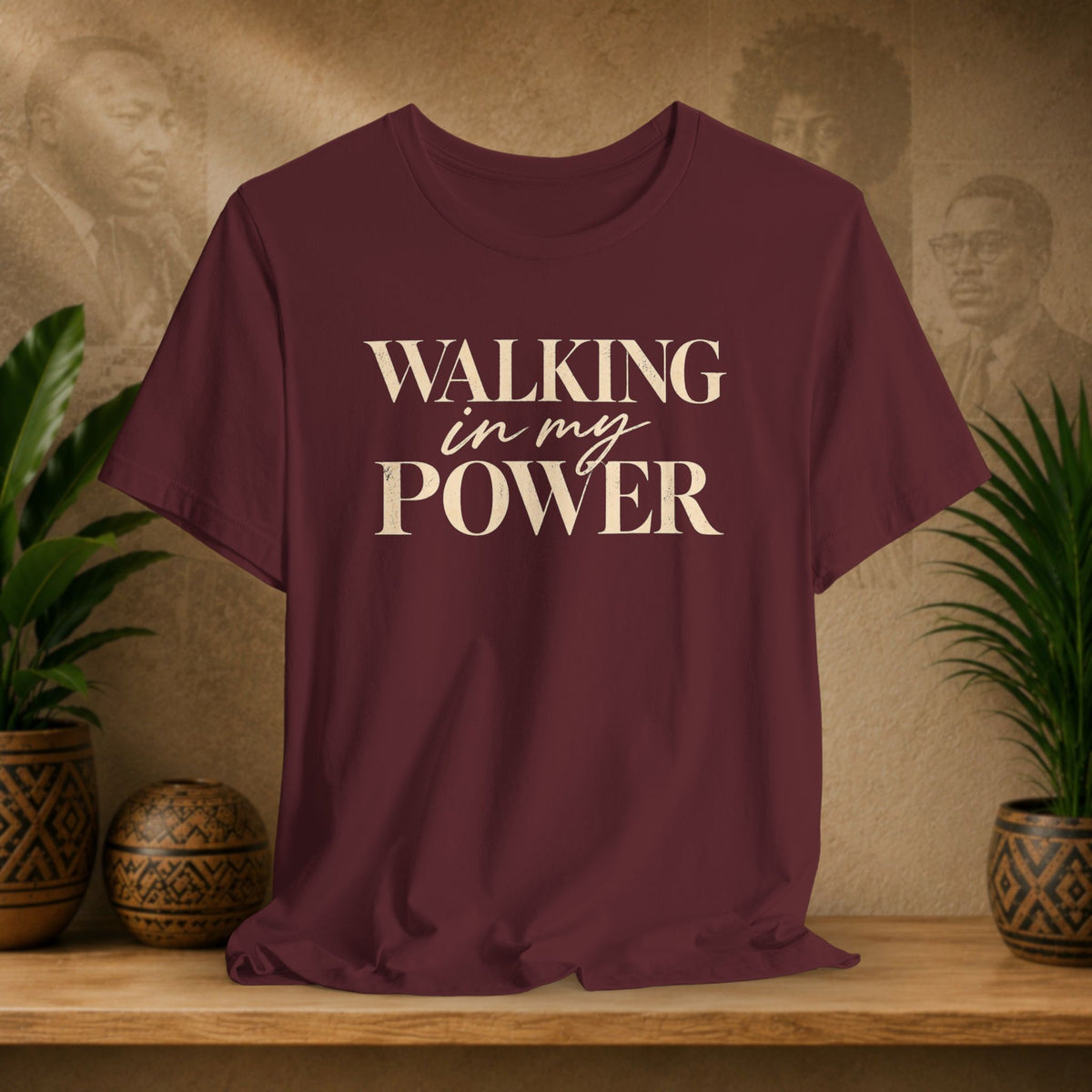 A maroon short-sleeve t-shirt with the text 'WALKING in my POWER' printed in beige on the front, displayed on a wooden shelf with decorative plants and a textured background.