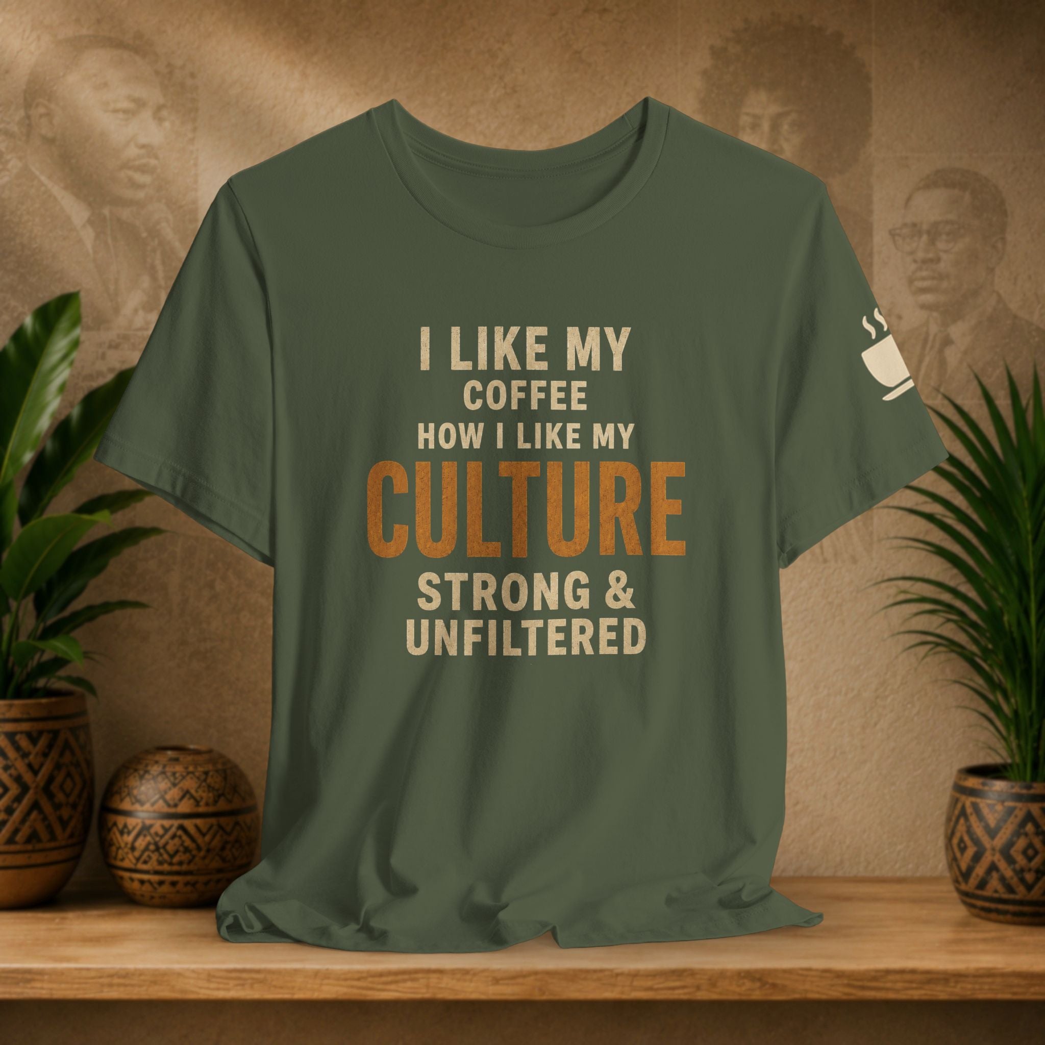 An olive green graphic t-shirt displayed on a wooden shelf, flanked by potted plants. The shirt features the text 'I LIKE MY COFFEE HOW I LIKE MY CULTURE STRONG & UNFILTERED' in cream and orange lettering, with a small white coffee cup graphic on the sleeve.