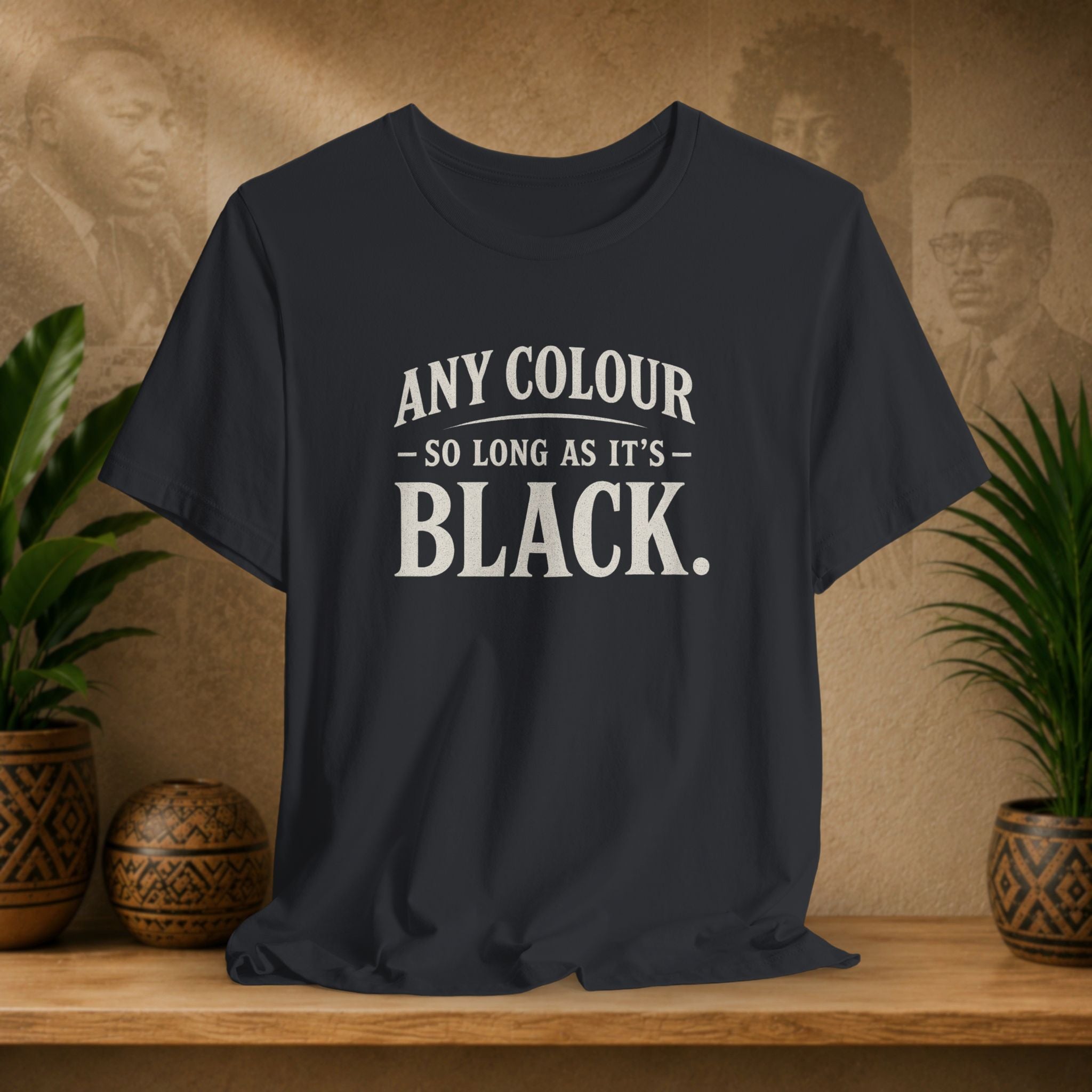 Black t-shirt with 'Any Colour So Long As It's Black' text on a wooden surface with plants and a textured wall background.