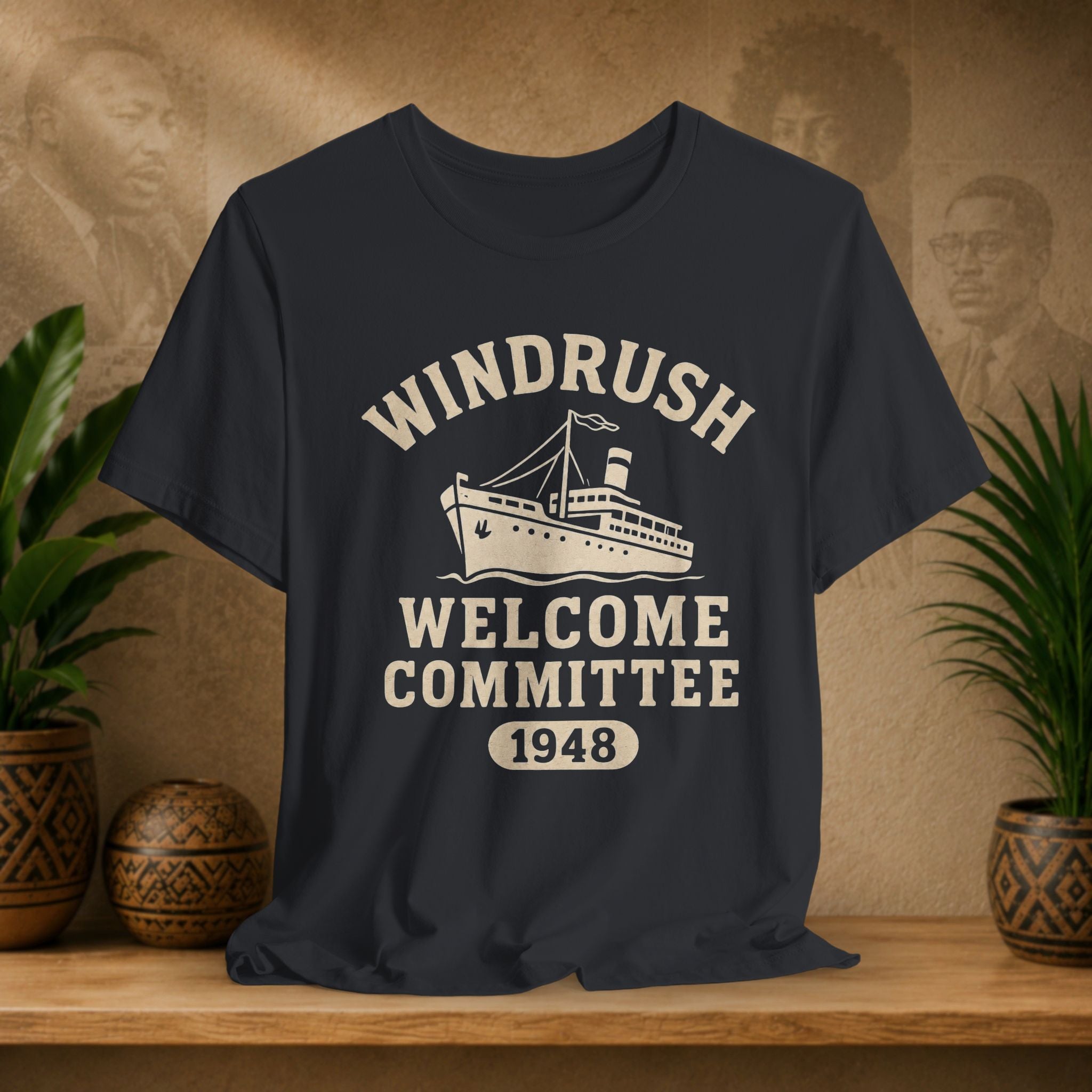 Black t-shirt with 'Windrush Welcome Committee 1948' design on a wooden surface with plants and a decorative wall.