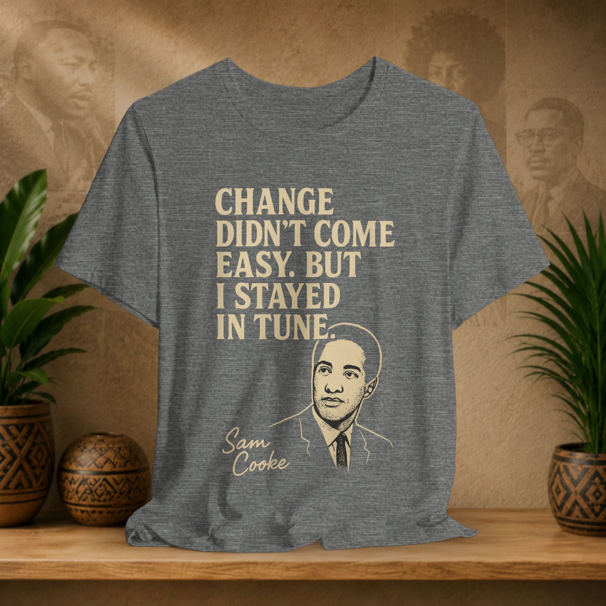 Gray t-shirt with a quote and portrait of Sam Cooke on a wooden surface with plants and a wall with portraits in the background.